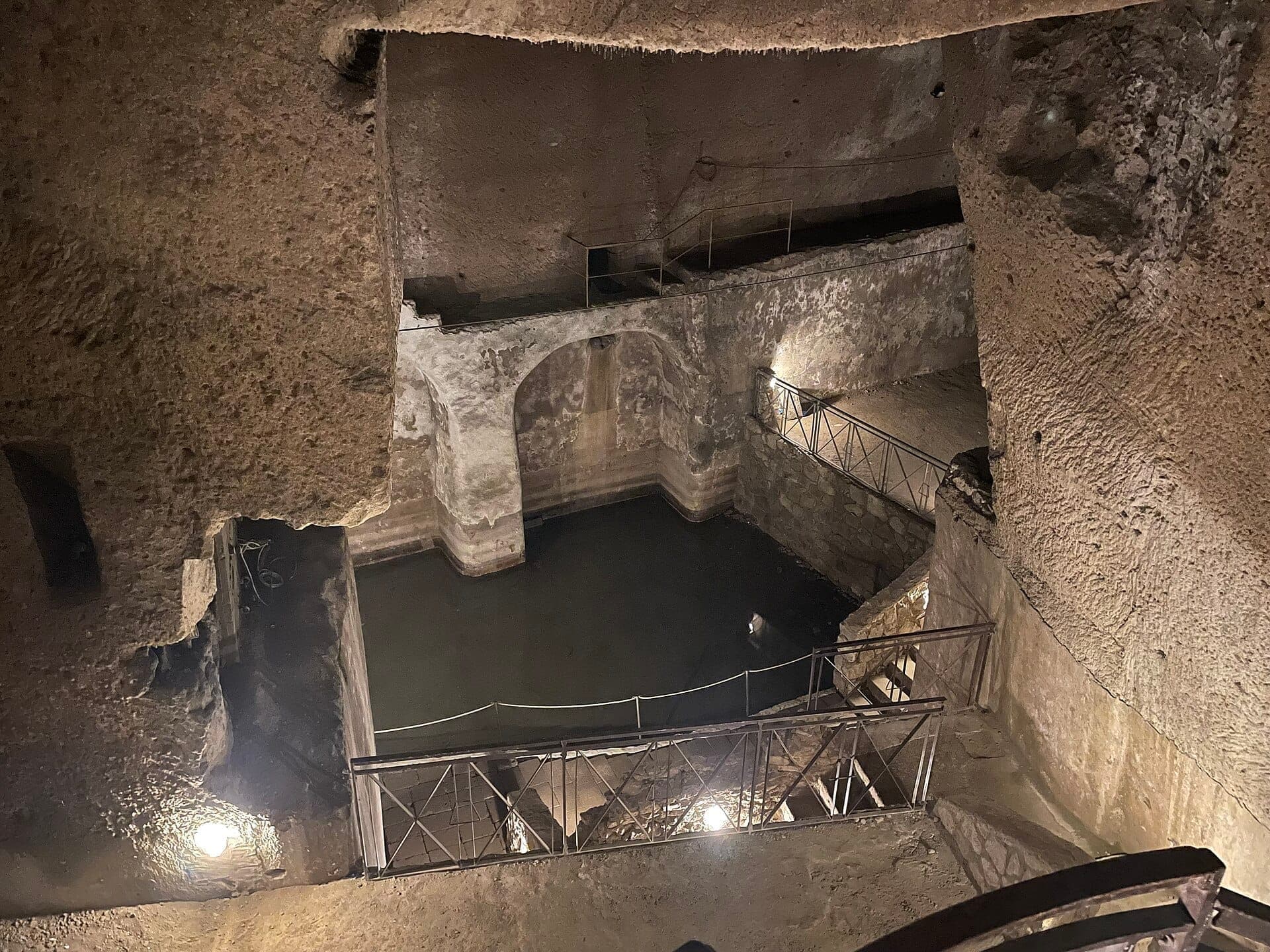 Vista dos antigos túneis de pedra e do reservatório de água subterrâneo de Napoli Sotterranea, iluminados por luzes suaves e rodeados por passarelas.