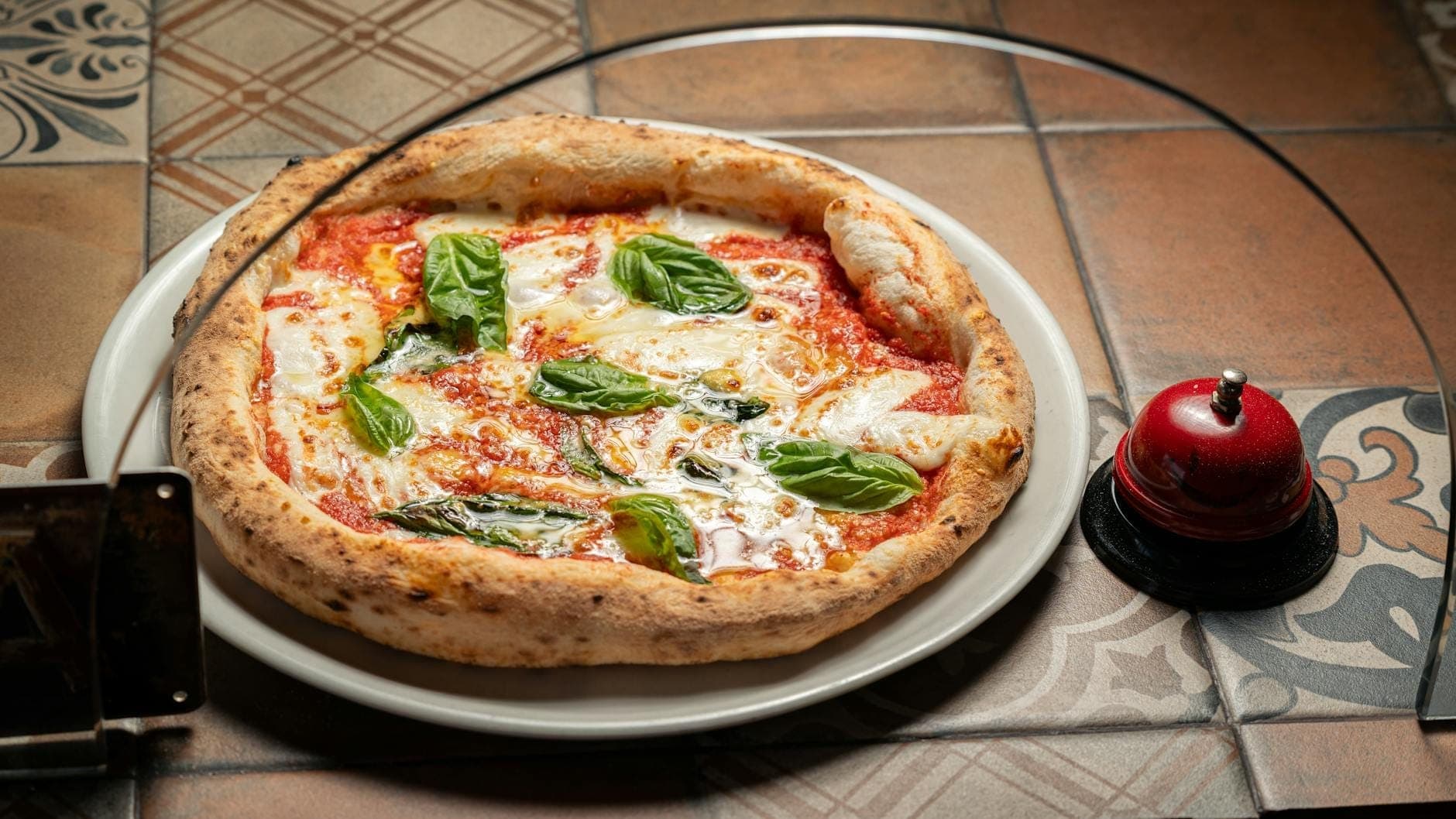 Pizza napolitana clássica com manjericão, molho de tomate e mozzarella em um prato branco sobre uma mesa de azulejos, evocando a renomada cultura da pizza de Nápoles.