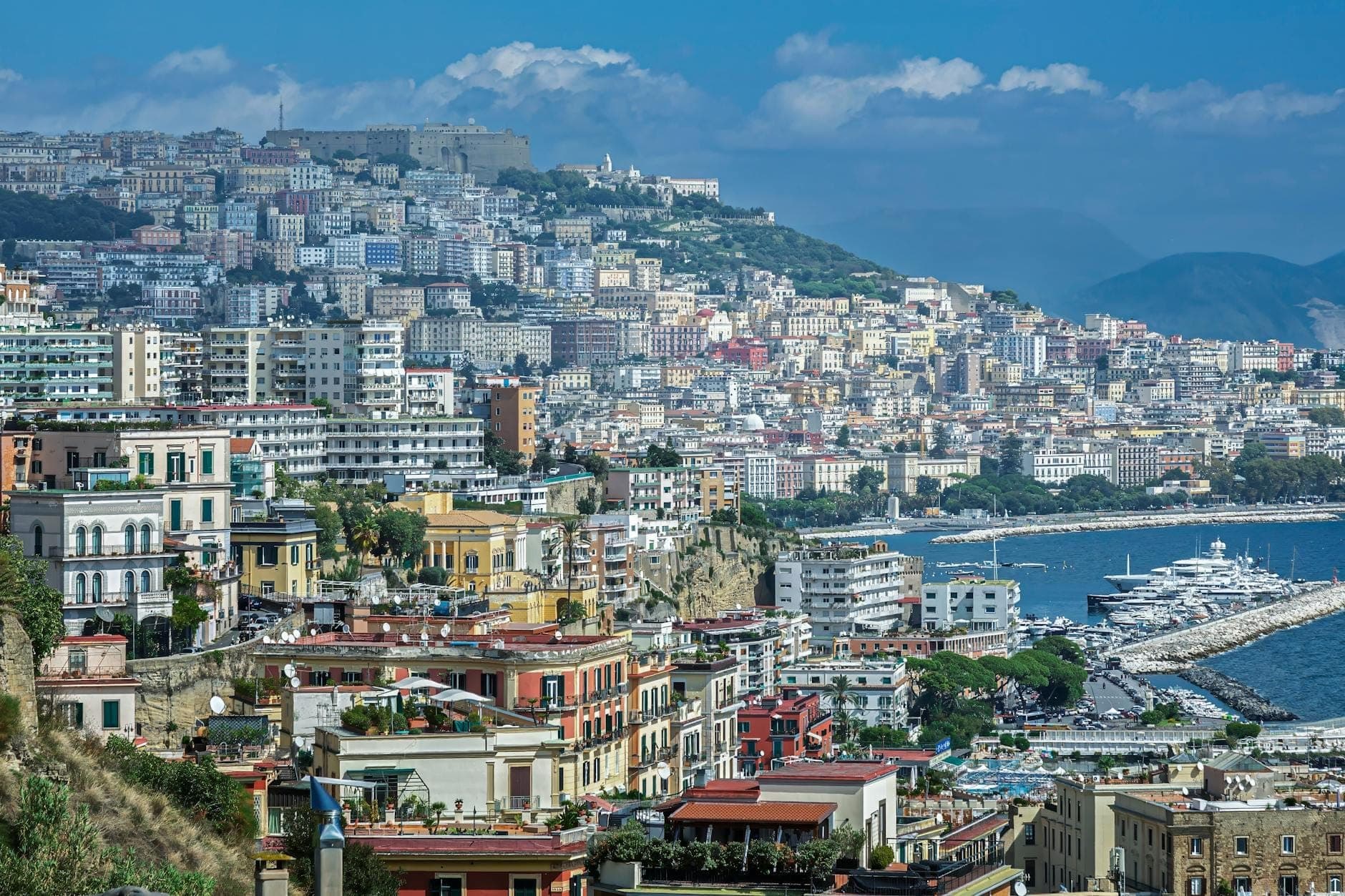 Vista panorâmica de Nápoles com edifícios coloridos, a marina à beira-mar e o Castel Sant'Elmo situado no topo de uma colina sob um céu azul.