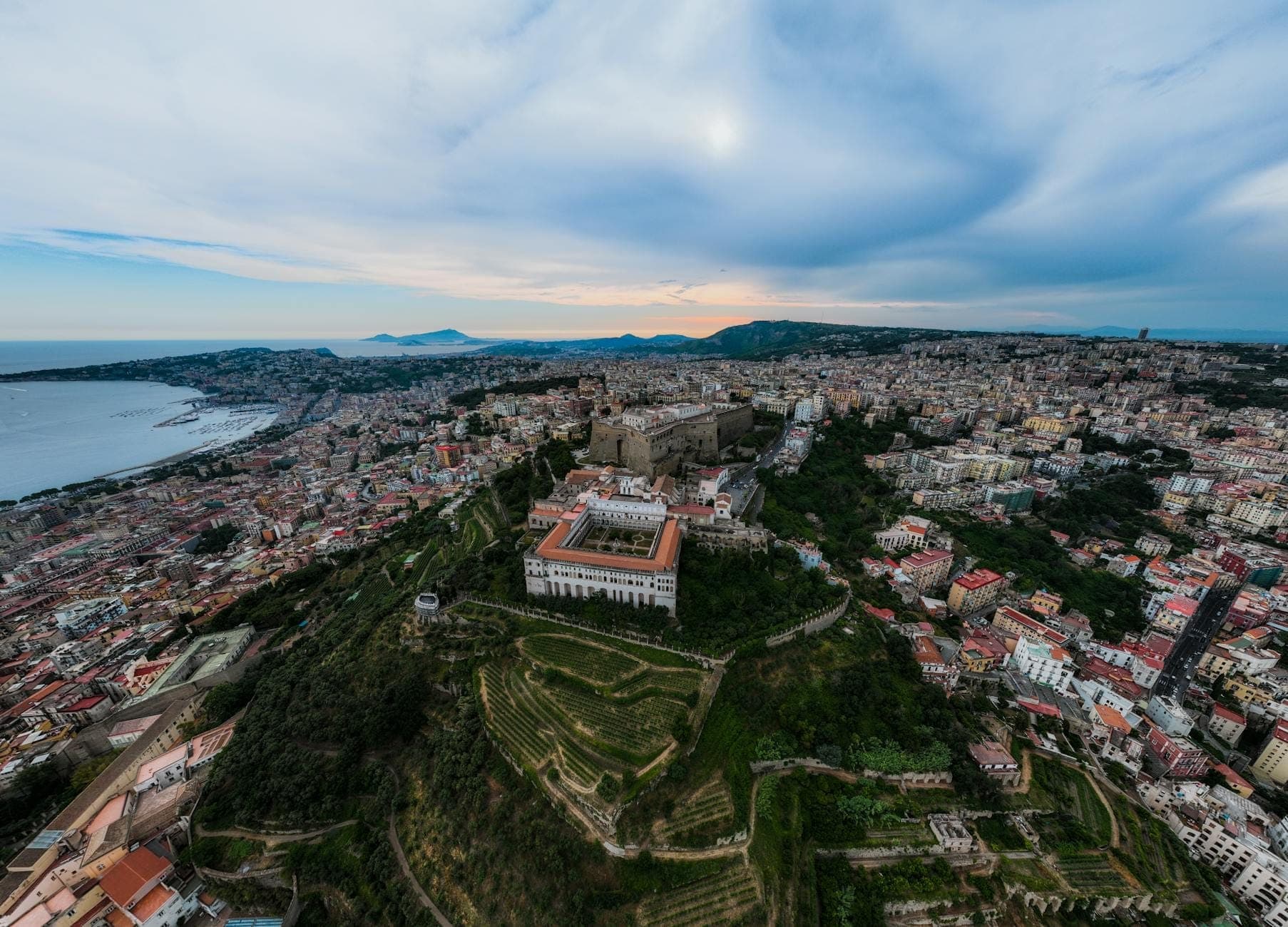 Vista aérea de Nápoles destacando o Vomero Hill com a Certosa di San Martino e a paisagem urbana que se estende em direção à costa sob um céu dramático.