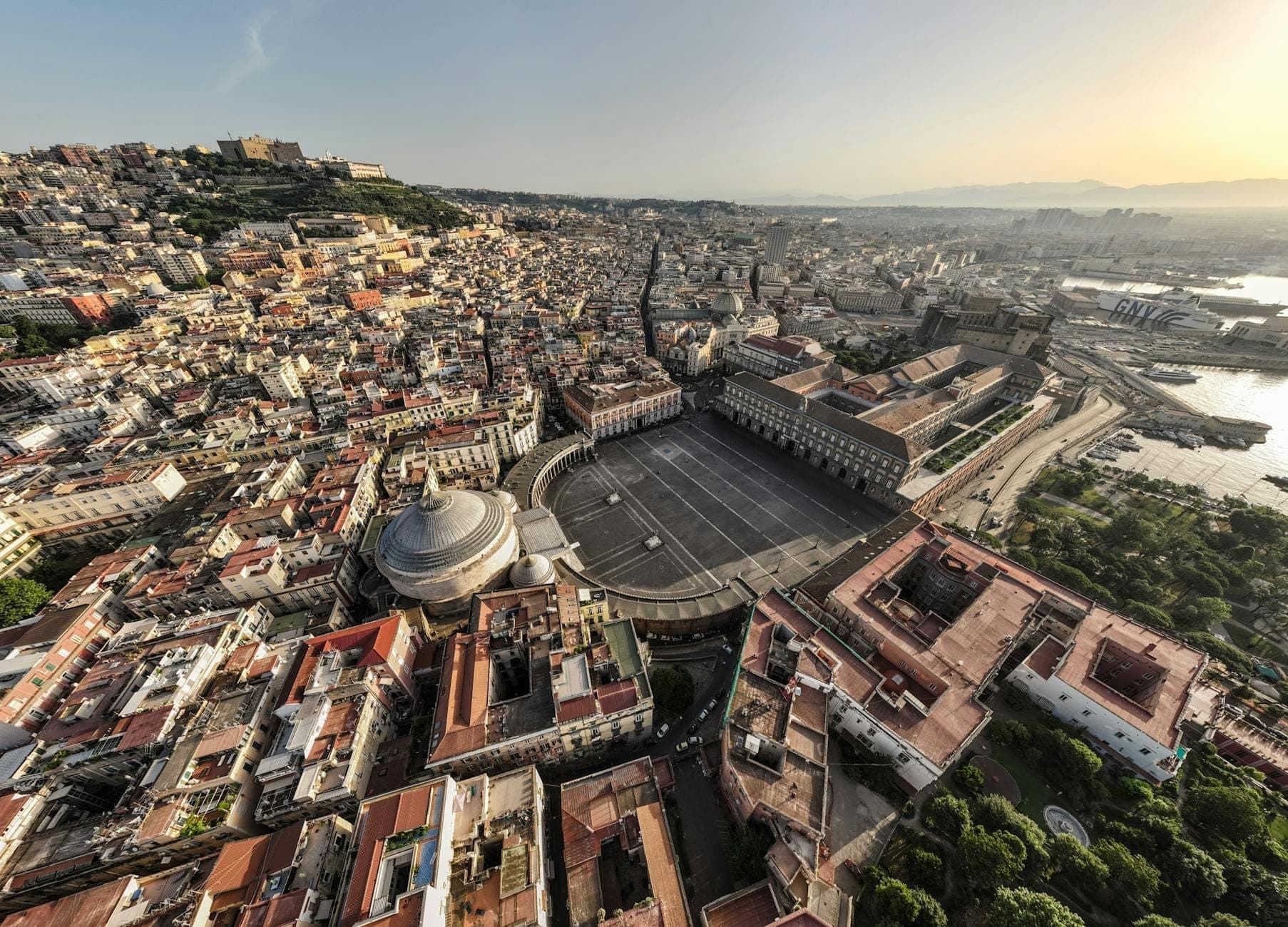 Vista aérea abrangente de Nápoles com a Piazza del Plebiscito, o Palazzo Reale, o Castel dell'Ovo e a baía ao pôr do sol.
