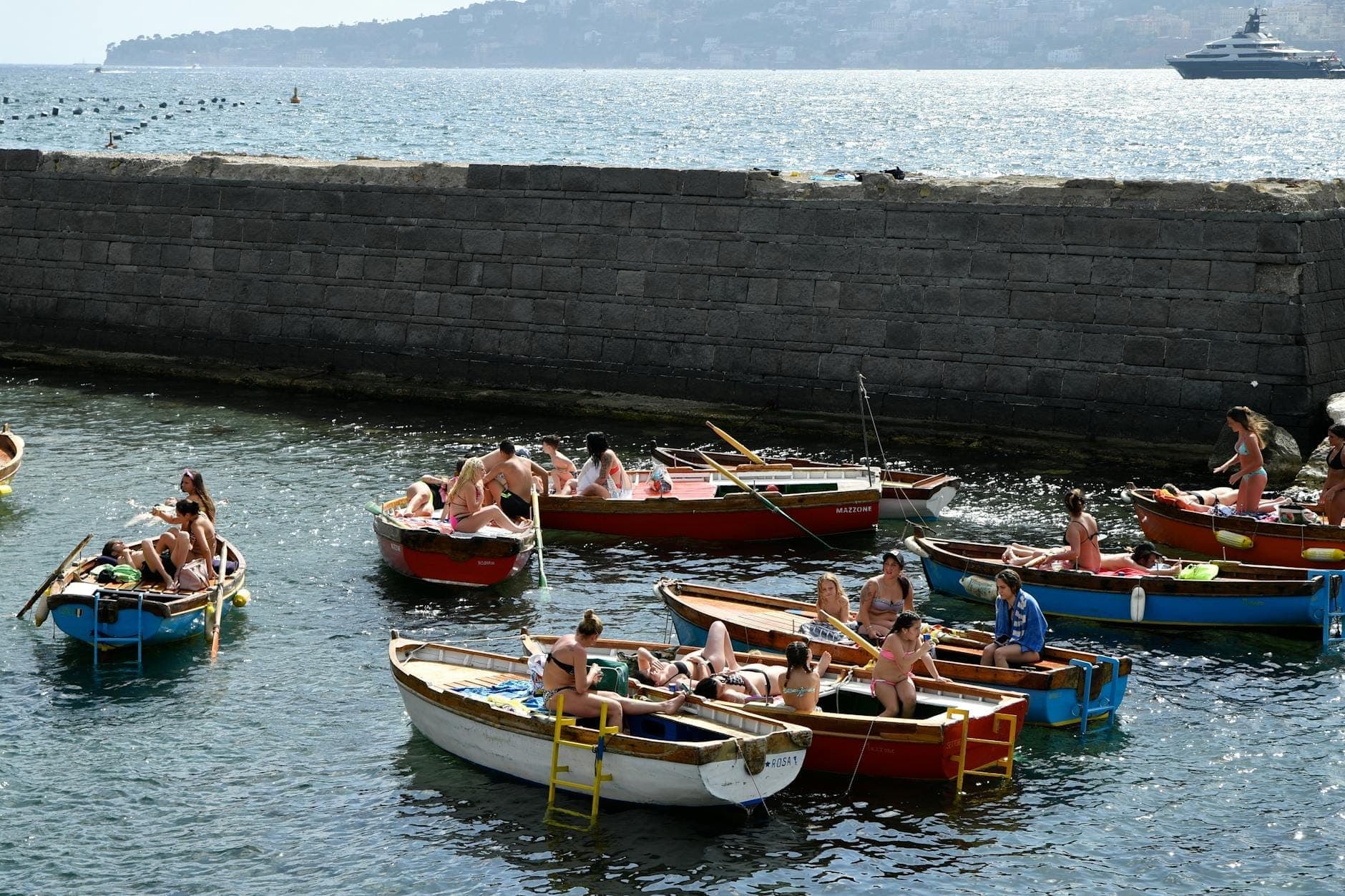 Famílias e amigos relaxam em barcos a remo de madeira coloridos perto de um molhe de pedra na Baía de Nápoles, com colinas distantes e iates visíveis.
