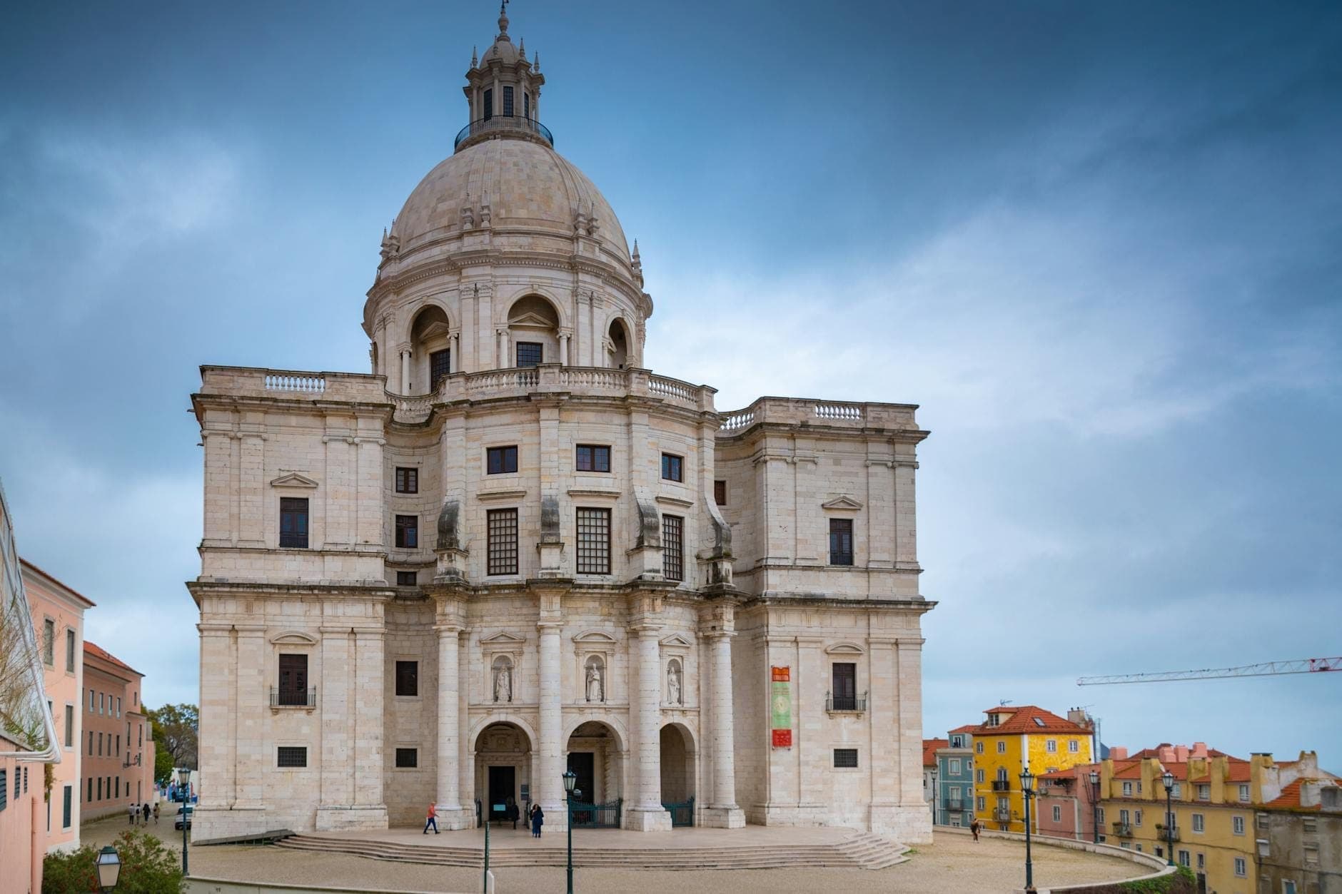 Vista frontal do Panteão Nacional em Lisboa sob um céu dramático, mostrando a fachada barroca e a icônica cúpula com casas coloridas ao redor.