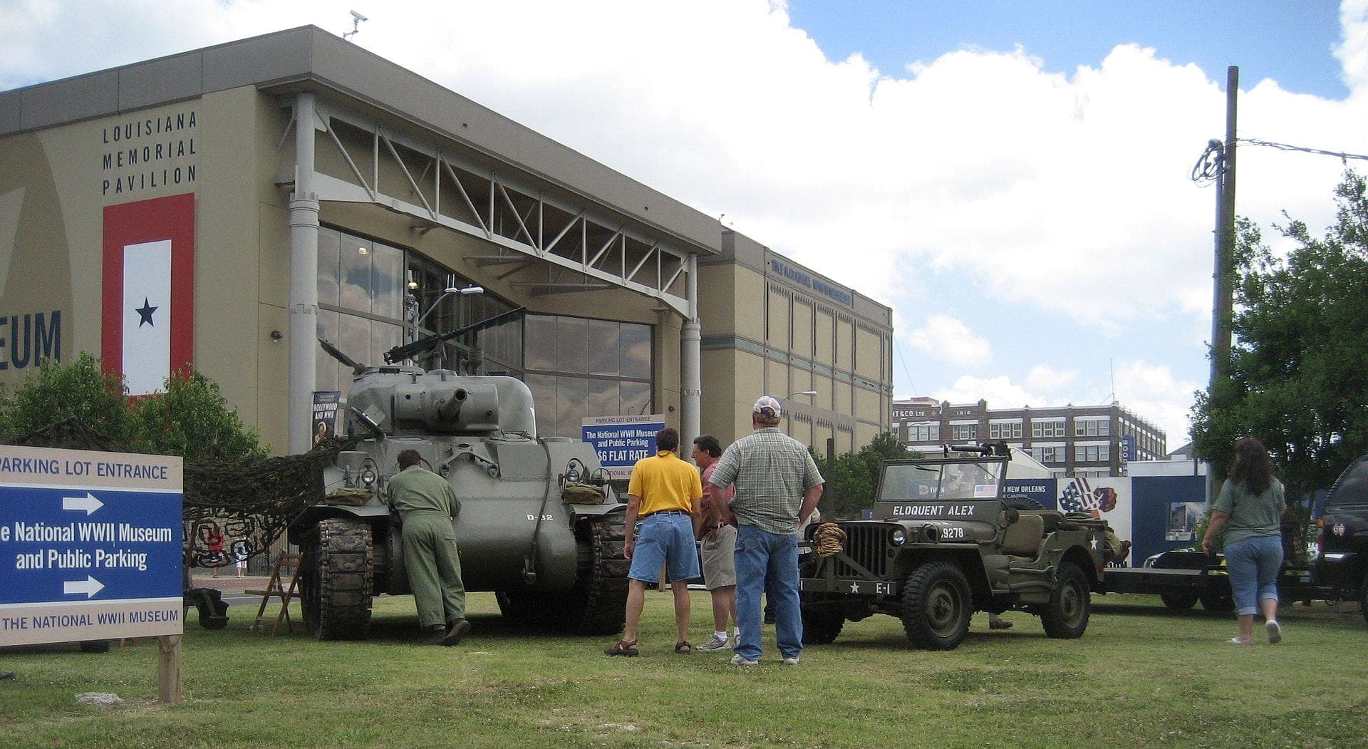 Visitors gather around a historic tank and jeep displayed outside the National WWII Museum’s Louisiana Memorial Pavilion on a sunny day in New Orleans.