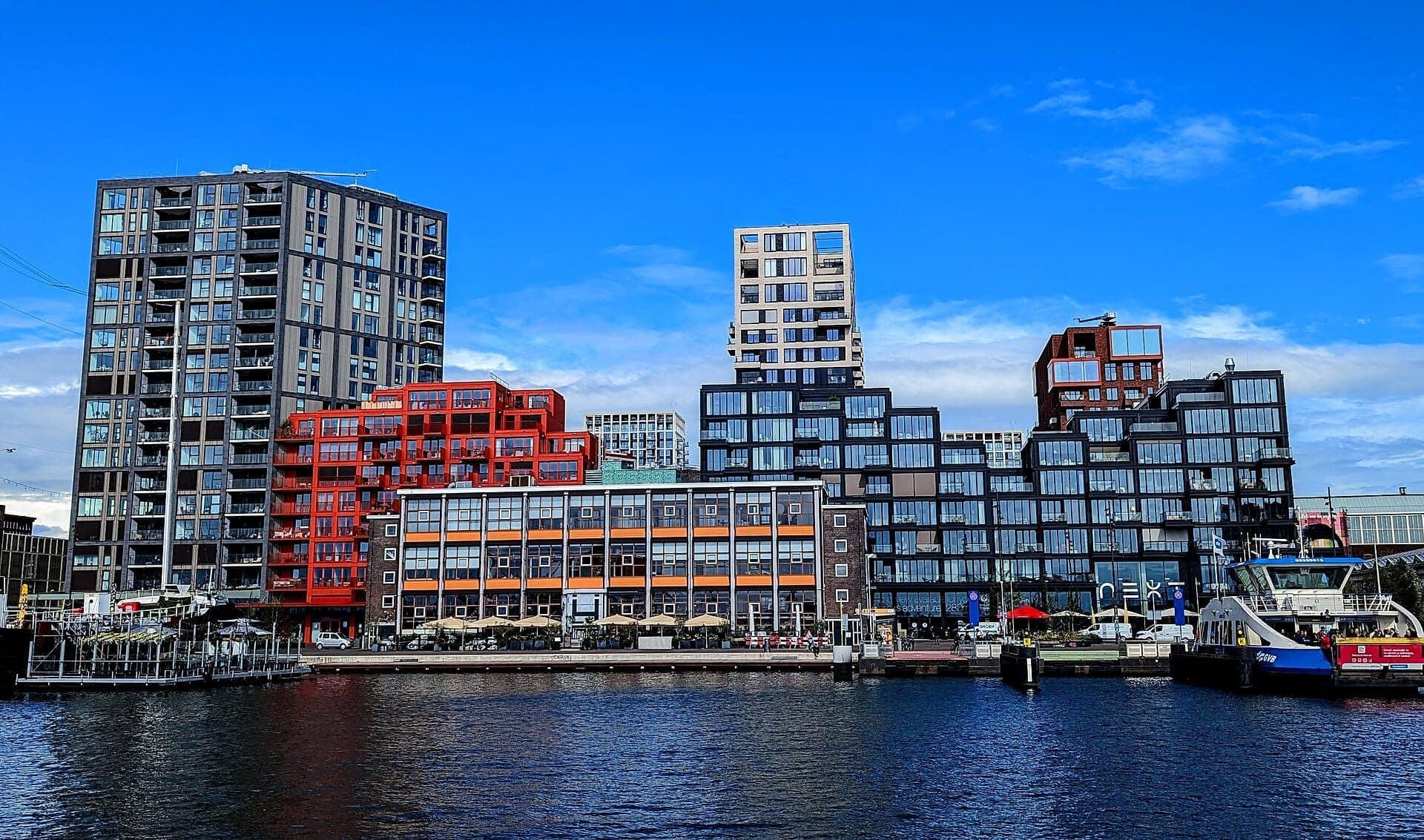 Moderne geometrische Gebäude des NDSM Wharf spiegeln sich im Fluss IJ unter einem strahlend blauen Himmel mit einer am Dock vertäuten Fähre.
