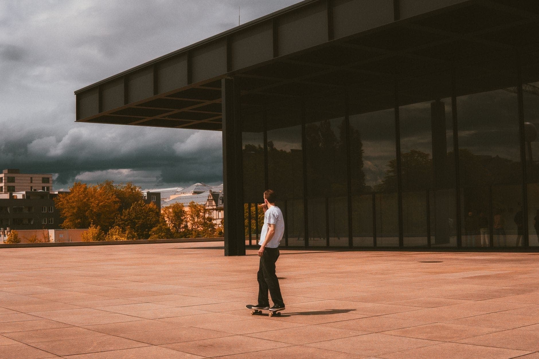 คนหนึ่งกำลังเล่นสเก็ตบอร์ดหน้า Neue Nationalgalerie เผยให้เห็นผนังกระจกและหลังคาเหล็กอันโดดเด่นใต้ท้องฟ้าดรามาติก