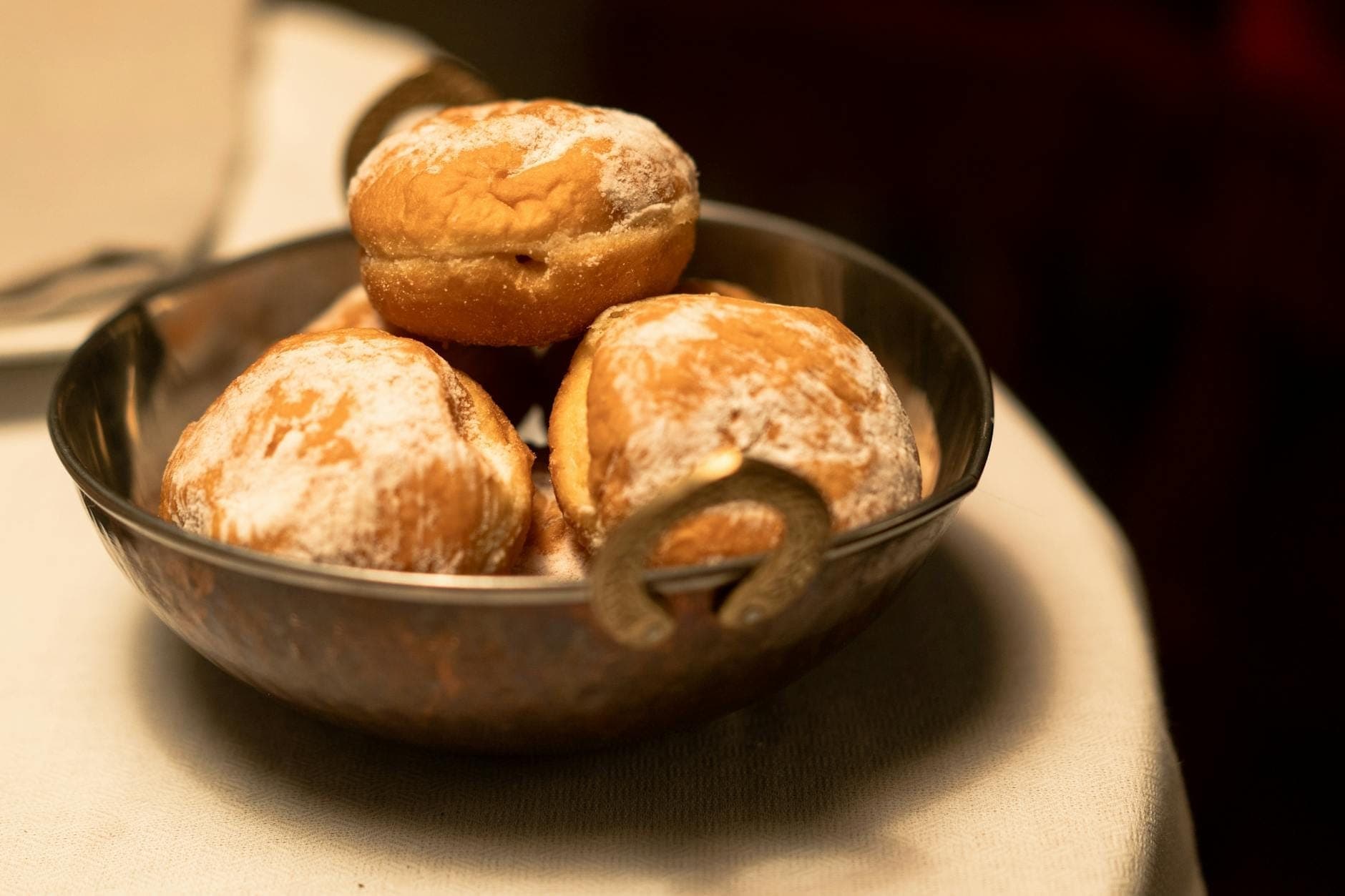 A close-up of golden beignets dusted with powdered sugar in a metal bowl on a white tablecloth, softly lit indoors.