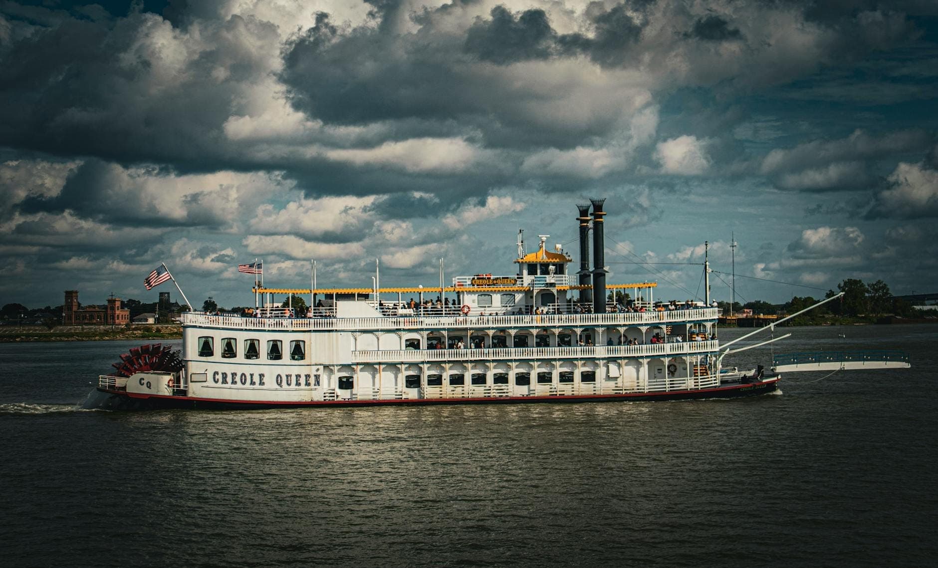 Um clássico barco a vapor de pás de Nova Orleans, o Creole Queen, navegando pelo rio Mississippi sob um céu nublado dramático.