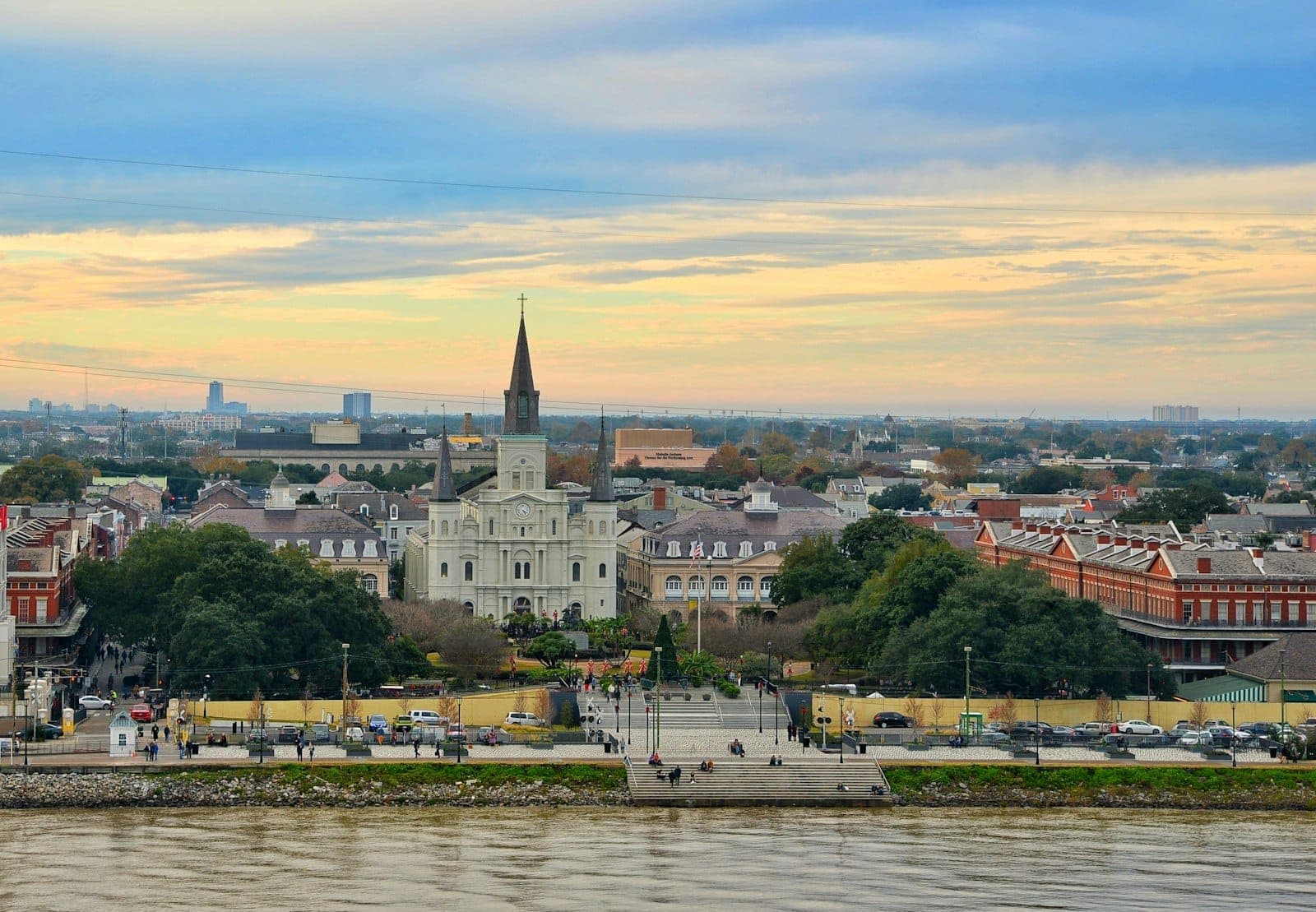 Iconic view of New Orleans featuring St. Louis Cathedral, Jackson Square, and historic French Quarter buildings under a colorful evening sky by the river.