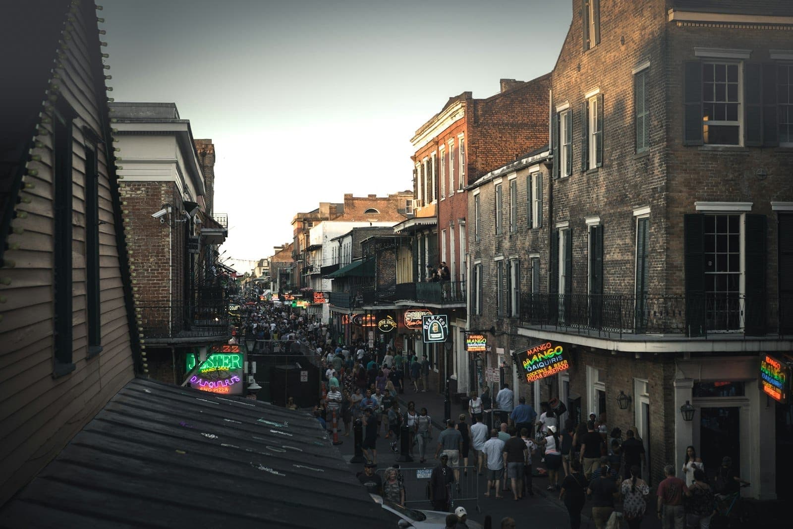 Crowds walk along Bourbon Street in New Orleans’ French Quarter, surrounded by historic brick buildings and bright neon signs at dusk.