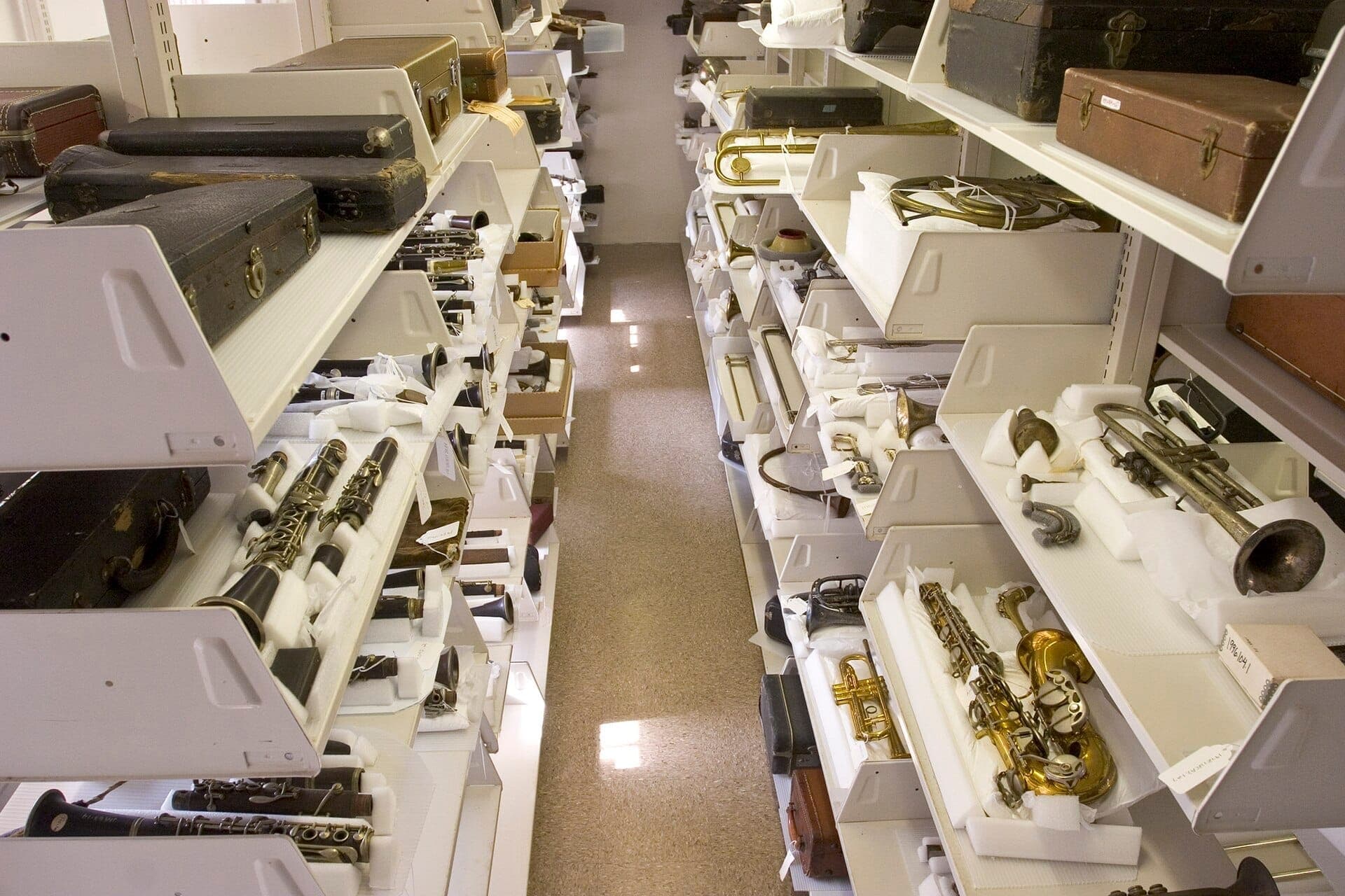 Rows of white shelves at the New Orleans Jazz Museum filled with vintage brass instruments, clarinets, and old instrument cases, under fluorescent lighting.
