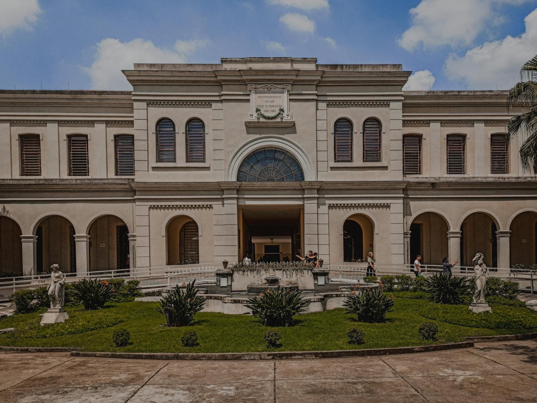 Gedung batu elegan dengan jendela lengkung dan halaman tengah yang menampilkan patung-patung dan taman hijau di bawah langit biru.