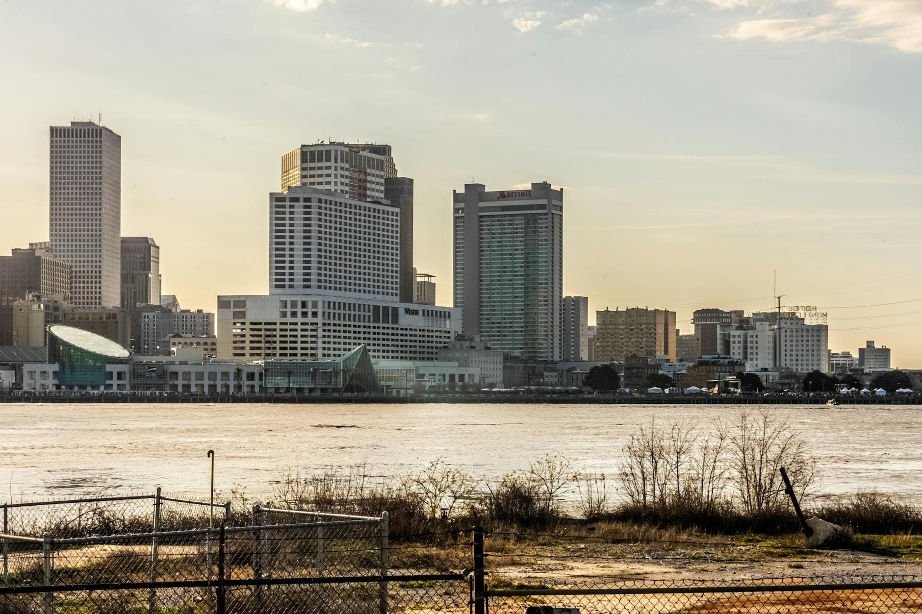 Pemandangan cakrawala pusat kota New Orleans dari seberang Sungai Mississippi dengan latar depan tepi sungai alami dan gedung-gedung kota di latar belakang.
