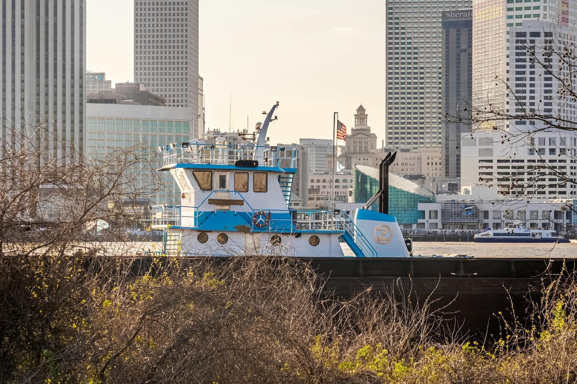 Sebuah kapal tunda melaju di sepanjang sungai dengan gedung pencakar langit pusat kota New Orleans di latar belakang, melambangkan aktivitas di tepi air.