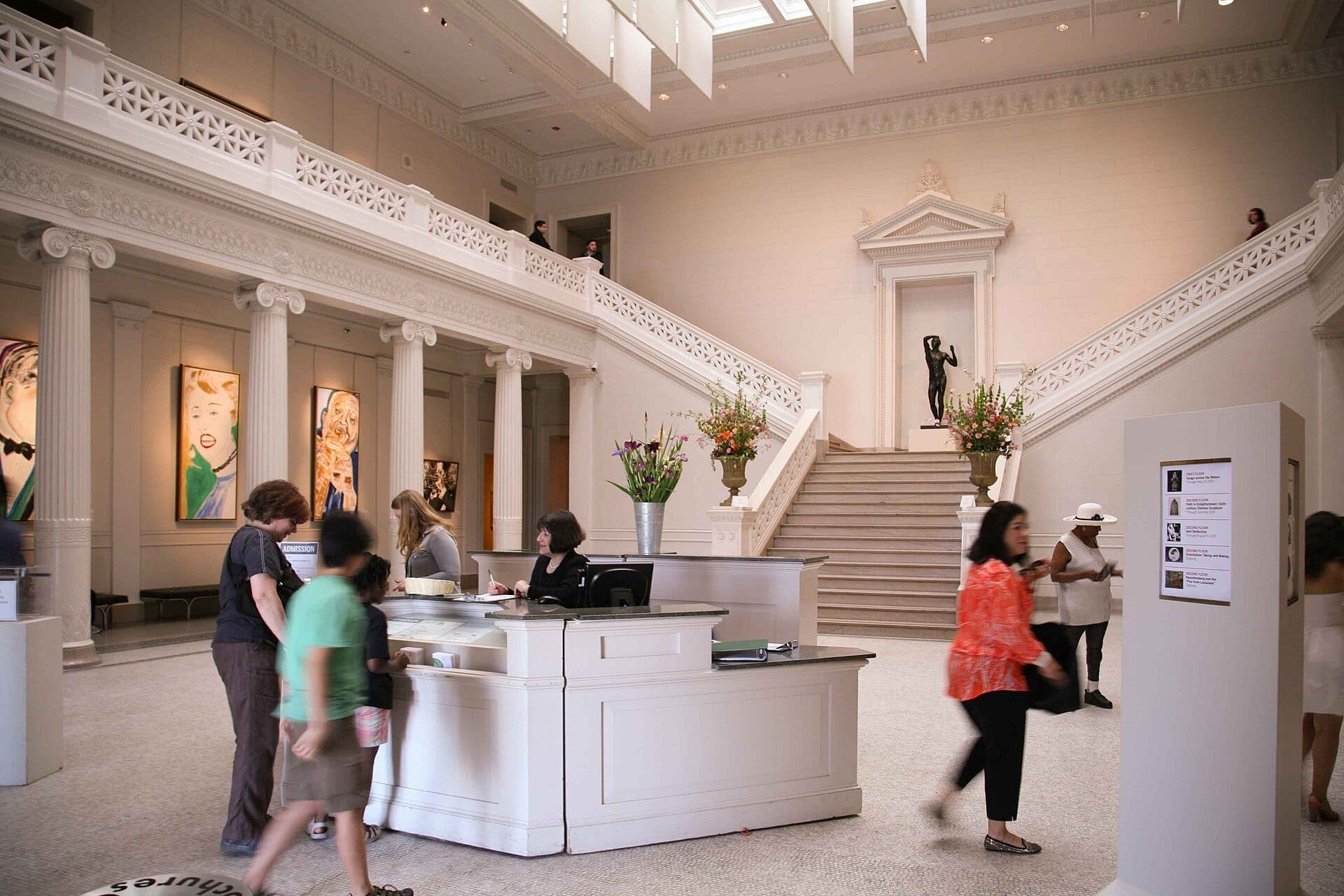 The grand entrance hall of the New Orleans Museum of Art with visitors, a reception desk, marble staircase, and classical architectural details.