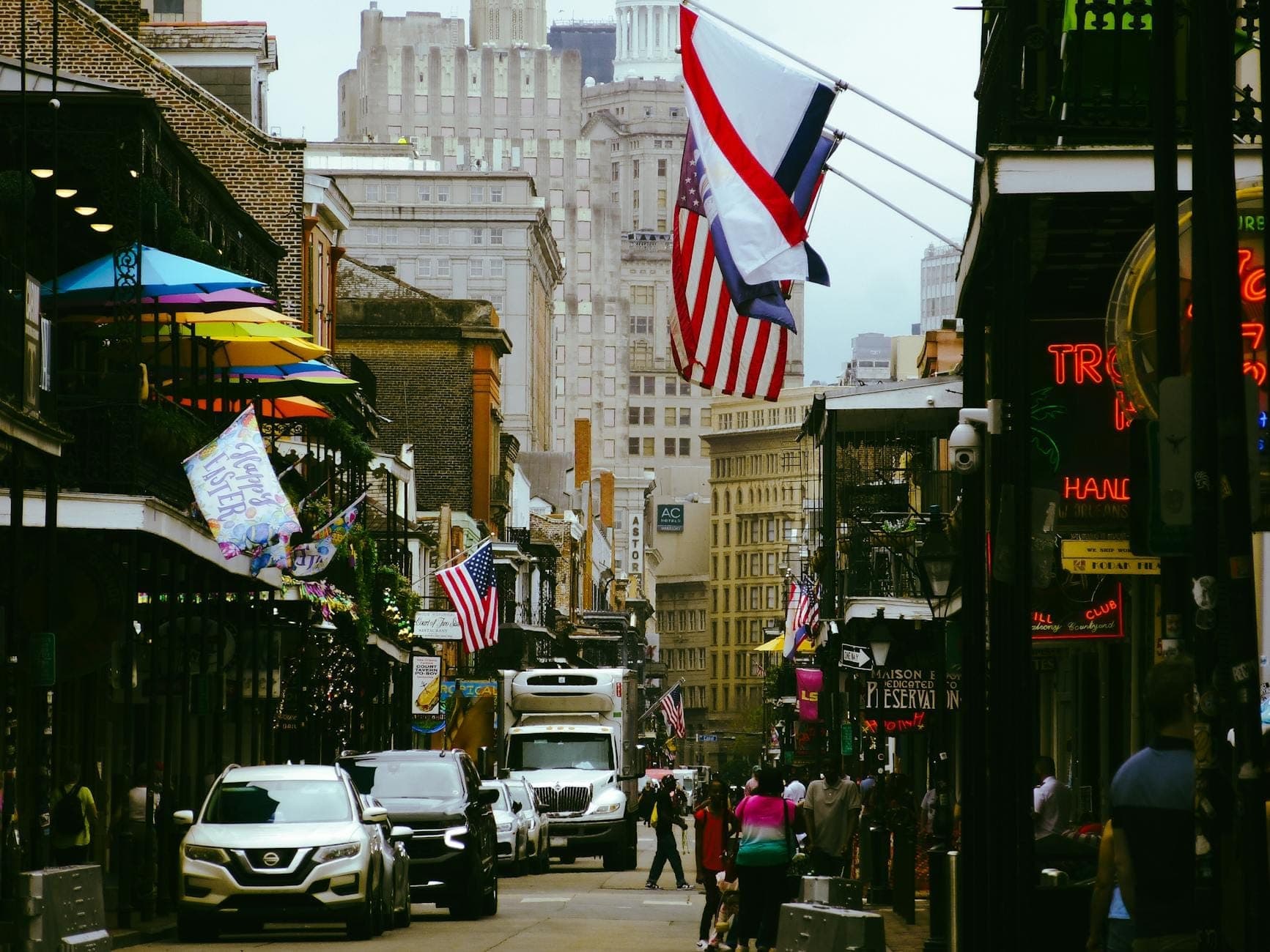 Bourbon Street im French Quarter mit amerikanischen Flaggen, bunten Balkonen, Menschenmengen und Autos bei Tag.