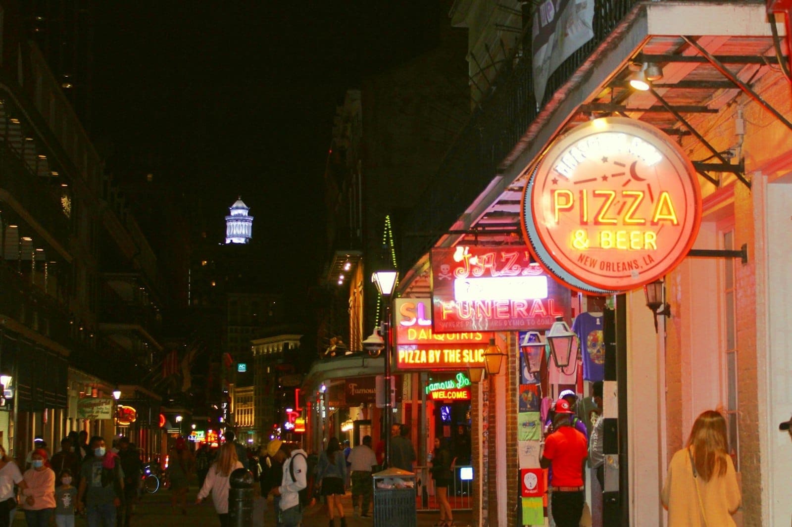 People walk along a lively New Orleans street at night, illuminated by neon bar and pizzeria signs and colorful city lights.