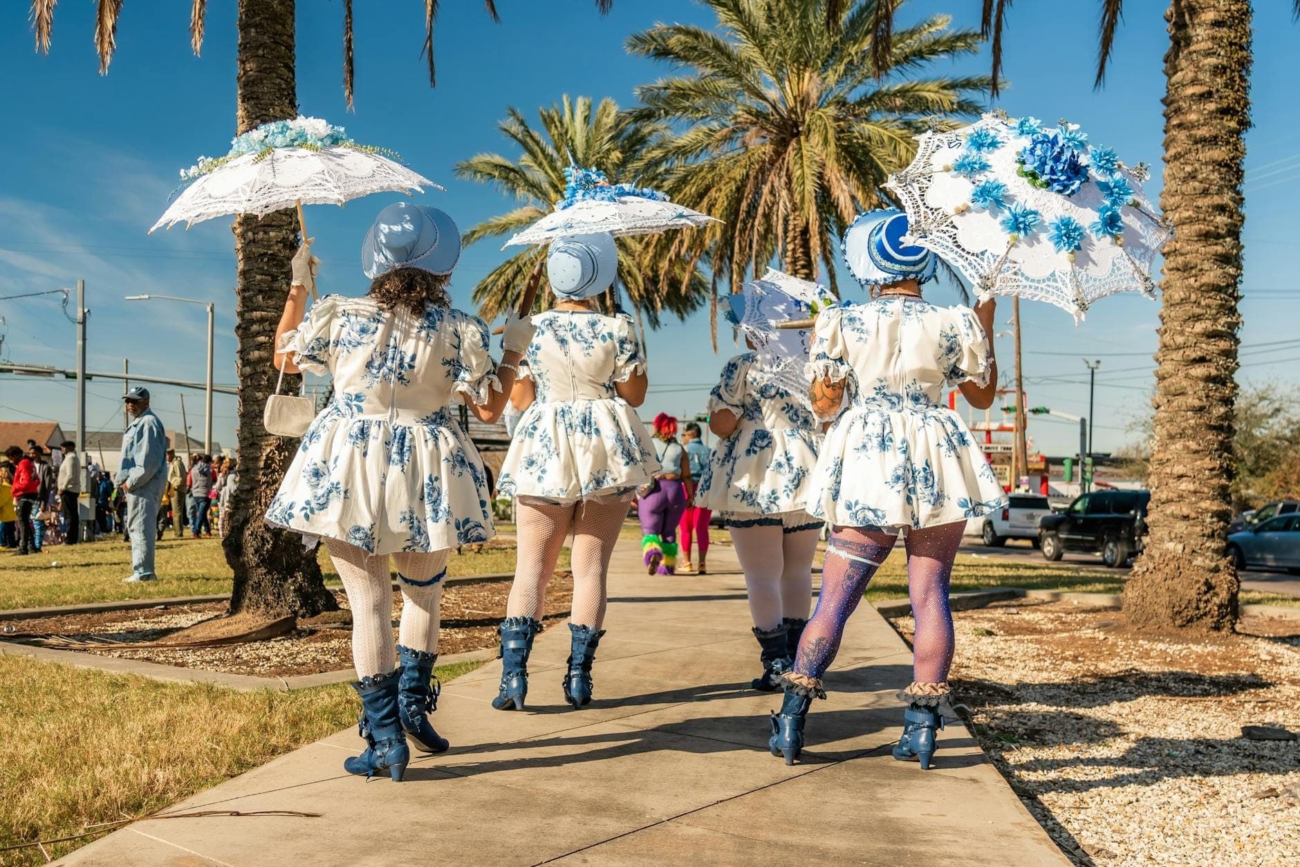Mulheres com trajes festivos e sombrinhas caminhando por uma rua de New Orleans durante um desfile ou evento importante, com palmeiras e céu azul.