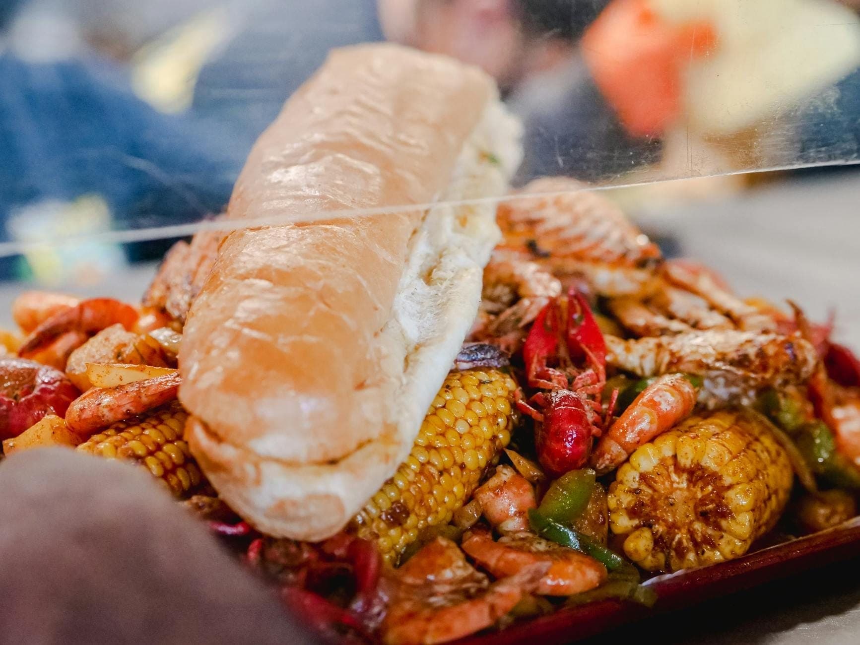 ภาพระยะใกล้ของจานซีฟู้ดต้ม New Orleans พร้อมกุ้งแม่น้ำ กุ้ง ข้าวโพด และแซนด์วิช po' boy วางอยู่ด้านบน