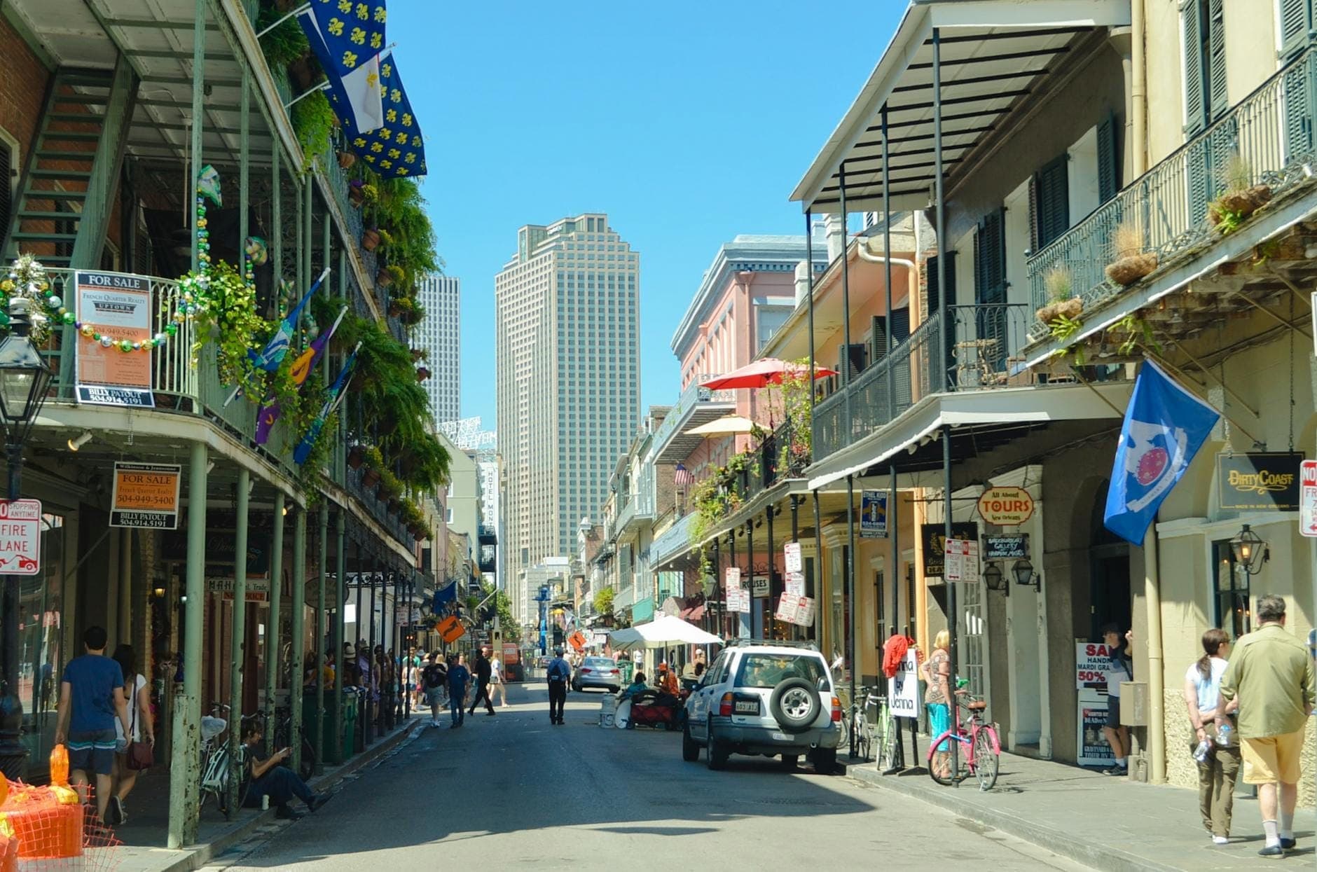 Cảnh ban ngày của một con phố sôi động ở French Quarter với những ban công sắt rèn cổ điển, cờ, người đi bộ và xe hơi tại New Orleans.
