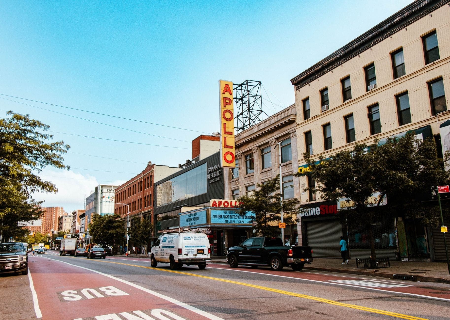 ภาพถนนหน้า Apollo Theater ที่มีป้ายไฟชื่อเสียงของฮาร์เล็ม รายล้อมด้วยตึกเก่าแก่และรถจอดริมถนนในวันที่แดดสดใส