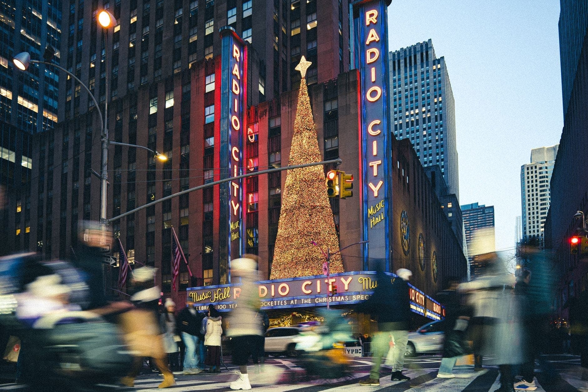 ฝูงชนเดินข้ามถนนกลางแมนฮัตตันที่เต็มไปด้วยสีสัน หน้าหอแสดง Radio City Music Hall และต้นคริสต์มาสขนาดยักษ์ที่สว่างไสวในบรรยากาศเทศกาล