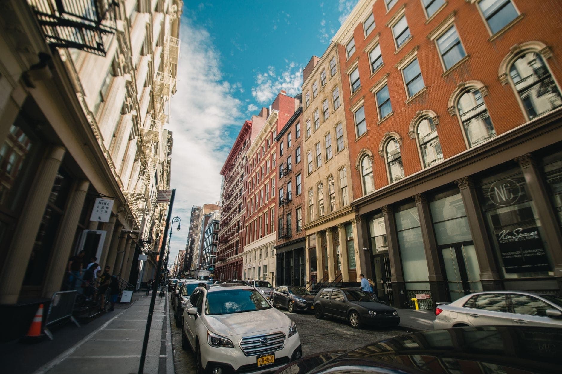 ภาพถนนคลาสสิกในย่าน SoHo กรุงนิวยอร์กซิตี้ พร้อมอาคารเหล็กหล่อ รถยนต์จอดเรียงรายตามถนนที่ปูด้วยหินกรวด และท้องฟ้าสีฟ้า