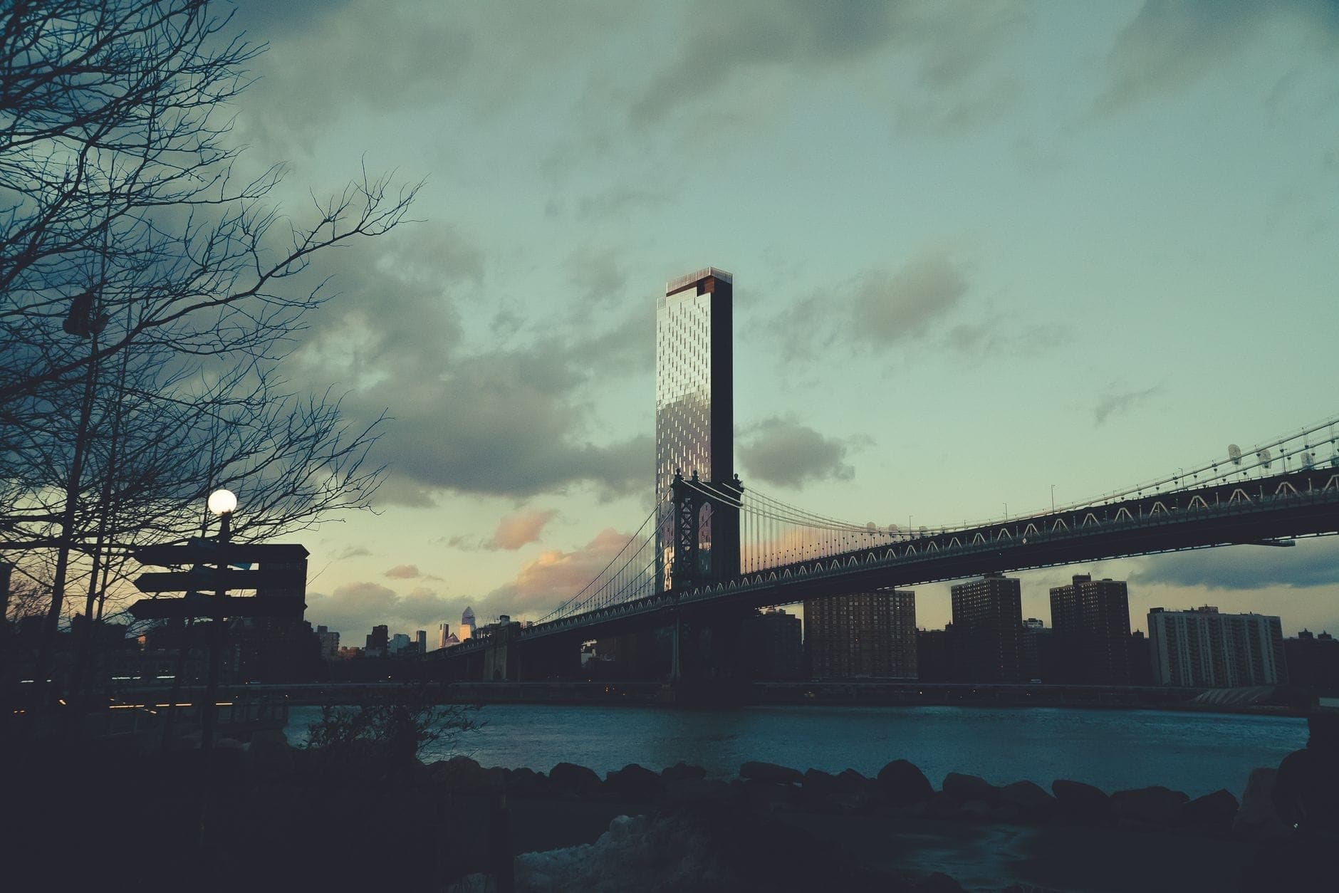 วิวของ Brooklyn Bridge ยามพระอาทิตย์ตกดิน พร้อมท้องฟ้าที่มีเมฆบางส่วนและเส้นขอบฟ้าของ New York City เป็นฉากหลัง