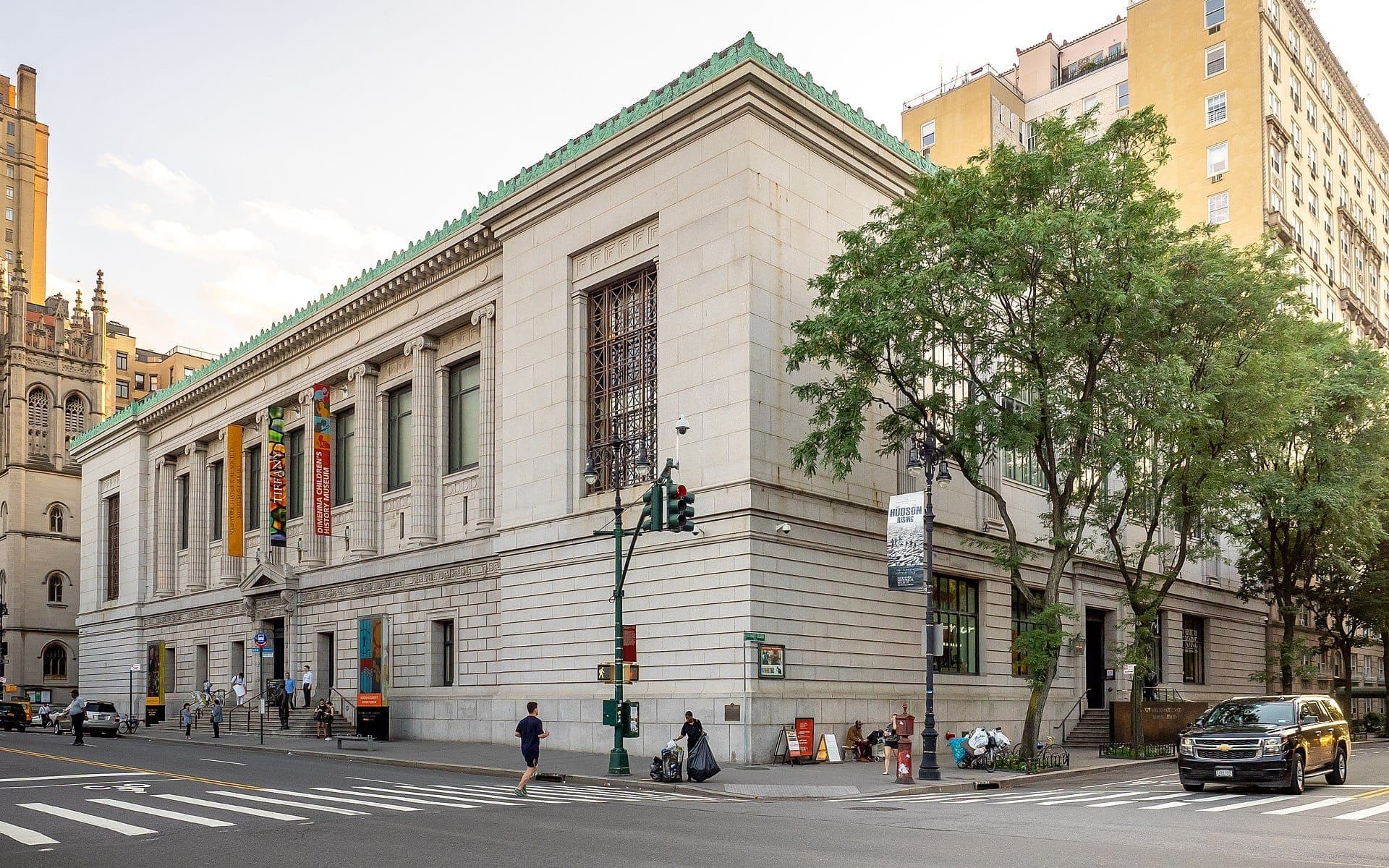 มุมมองด้านมุมของอาคารหินแบบนีโอคลาสสิกของ New-York Historical Society พร้อมทางเดินเรียงรายด้วยต้นไม้ ป้ายโปสเตอร์ของพิพิธภัณฑ์ และผู้คนที่เดินในวันที่แดดสดใส