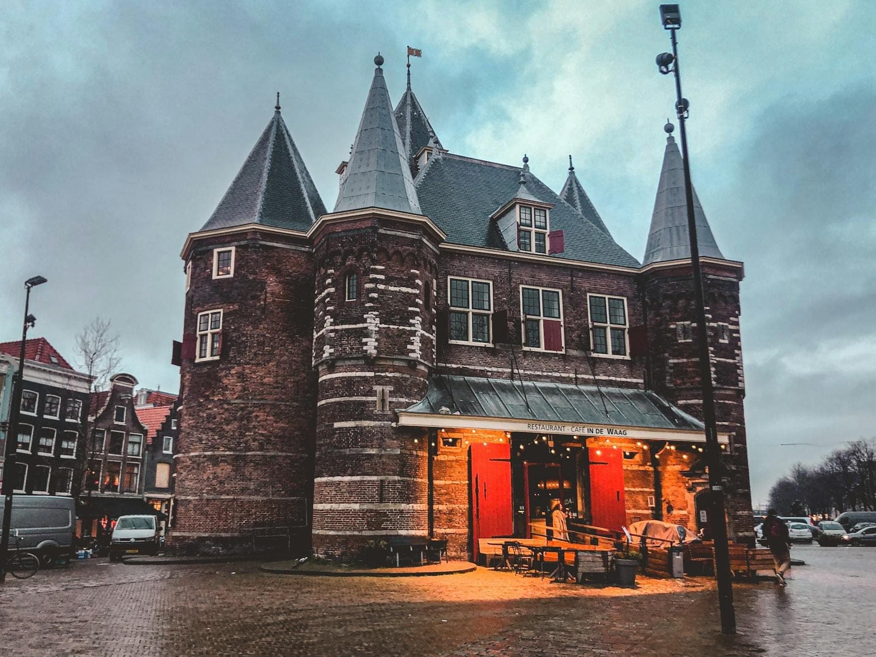Blick auf das historische De-Waag-Gebäude am Nieuwmarkt in Amsterdam in der Abenddämmerung, mit glühendem Licht und Kopfsteinpflaster davor.
