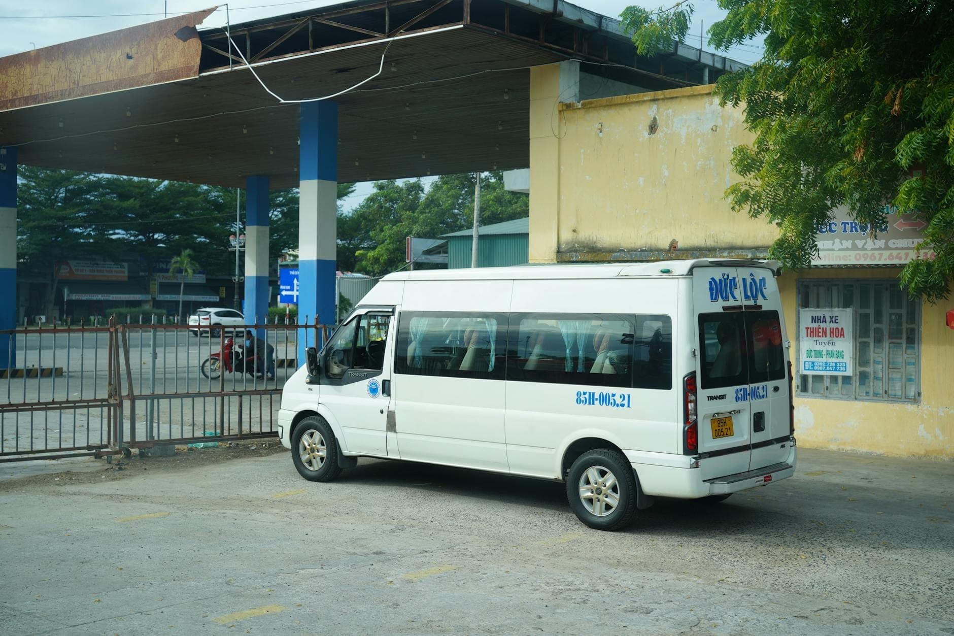Белый пассажирский микроавтобус, припаркованный на небольшом автобусном вокзале с жёлтой стеной и вывесками, типичный для региональных поездок по Вьетнаму.
