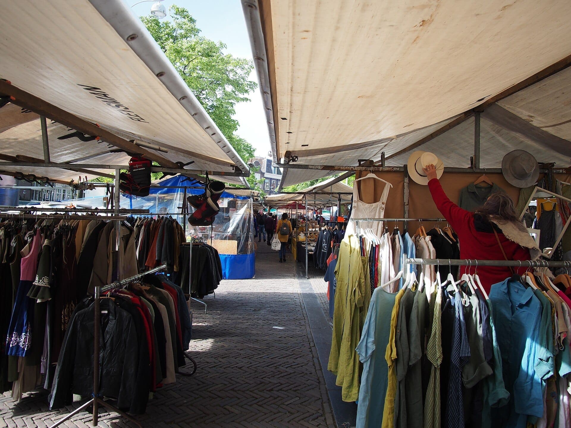 Stände voller bunter Kleidung, Jacken und Hüte säumen den belebten Noordermarkt-Platz in Amsterdam unter breiten, sonnenbeschienenen Markisen.