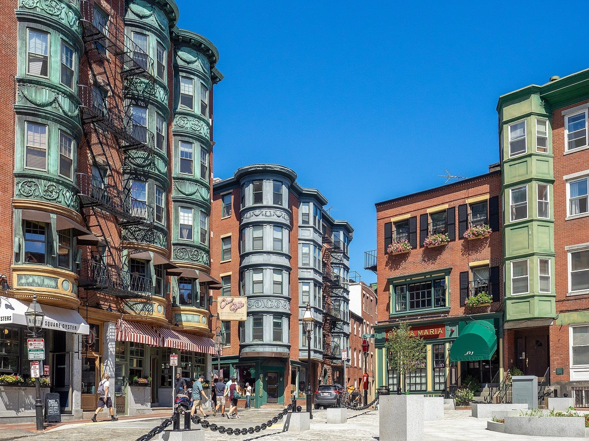 Des bâtiments historiques en briques et cuivre longent une rue ensoleillée dans le North End de Boston, avec des passants devant des cafés et restaurants sous un ciel bleu dégagé.