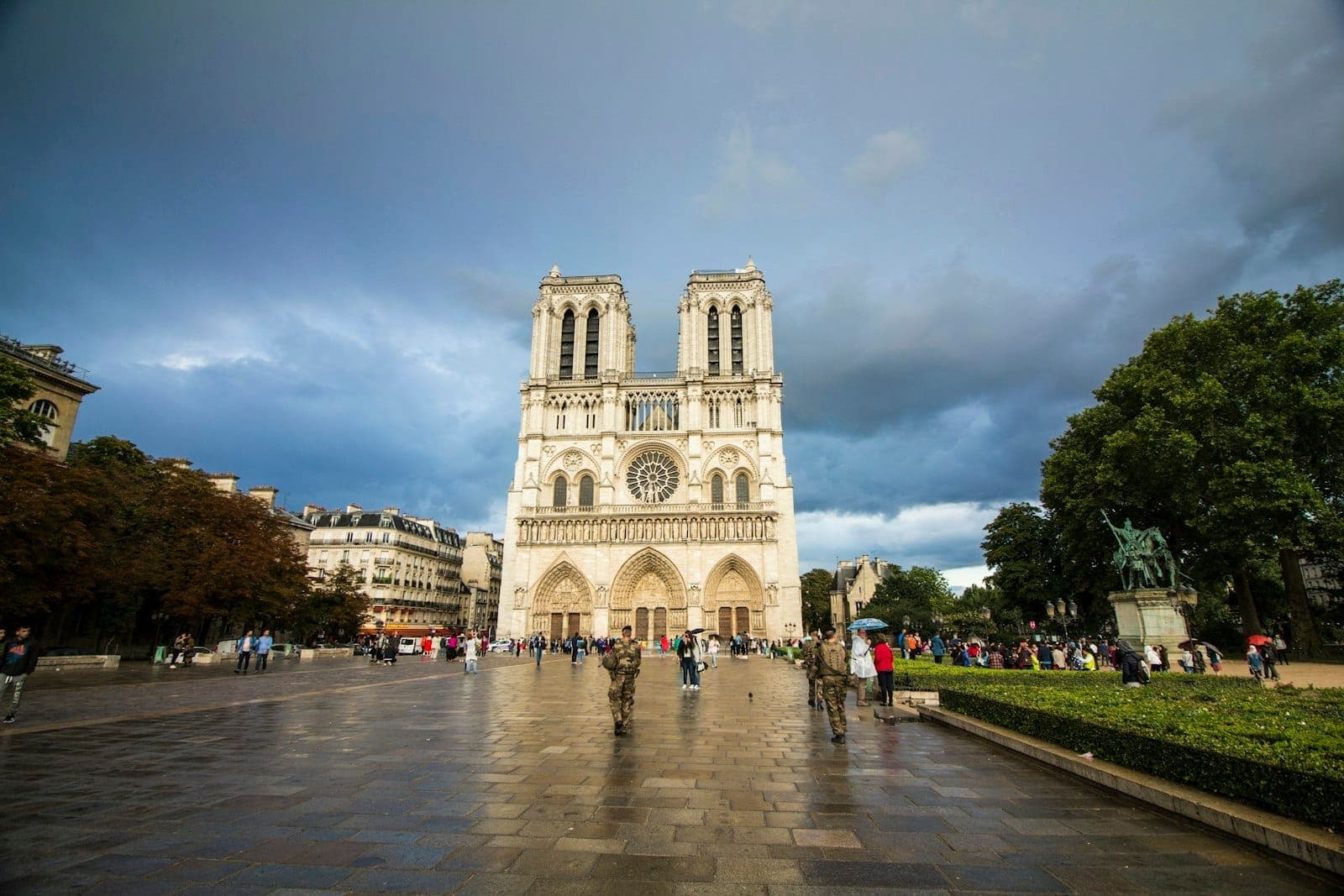 Wide view of Notre-Dame Cathedral’s iconic front facade with twin towers, cloudy sky, and visitors gathered in the main square, showcasing its stunning Gothic architecture.