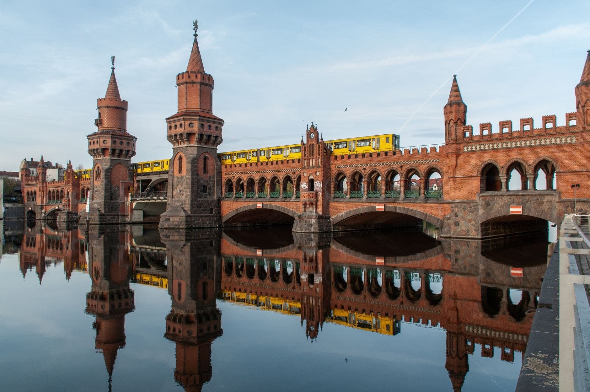 มุมกว้างของหอคอยและซุ้มอิฐนีโอโกธิค Oberbaumbrücke สะท้อนบนแม่น้ำ Spree สงบ ๆ พร้อมขบวนรถไฟ U-Bahn สีเหลืองแล่นข้ามสะพาน ภายใต้ท้องฟ้าสว่าง