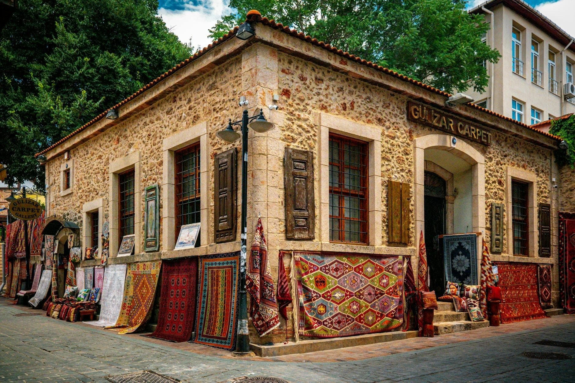 Ecke eines alten Steingebäudes im Basar-Viertel von Antalya, gesäumt von bunten Teppichen und Marktständen auf Kopfsteinpflaster, umgeben von historischer Architektur.