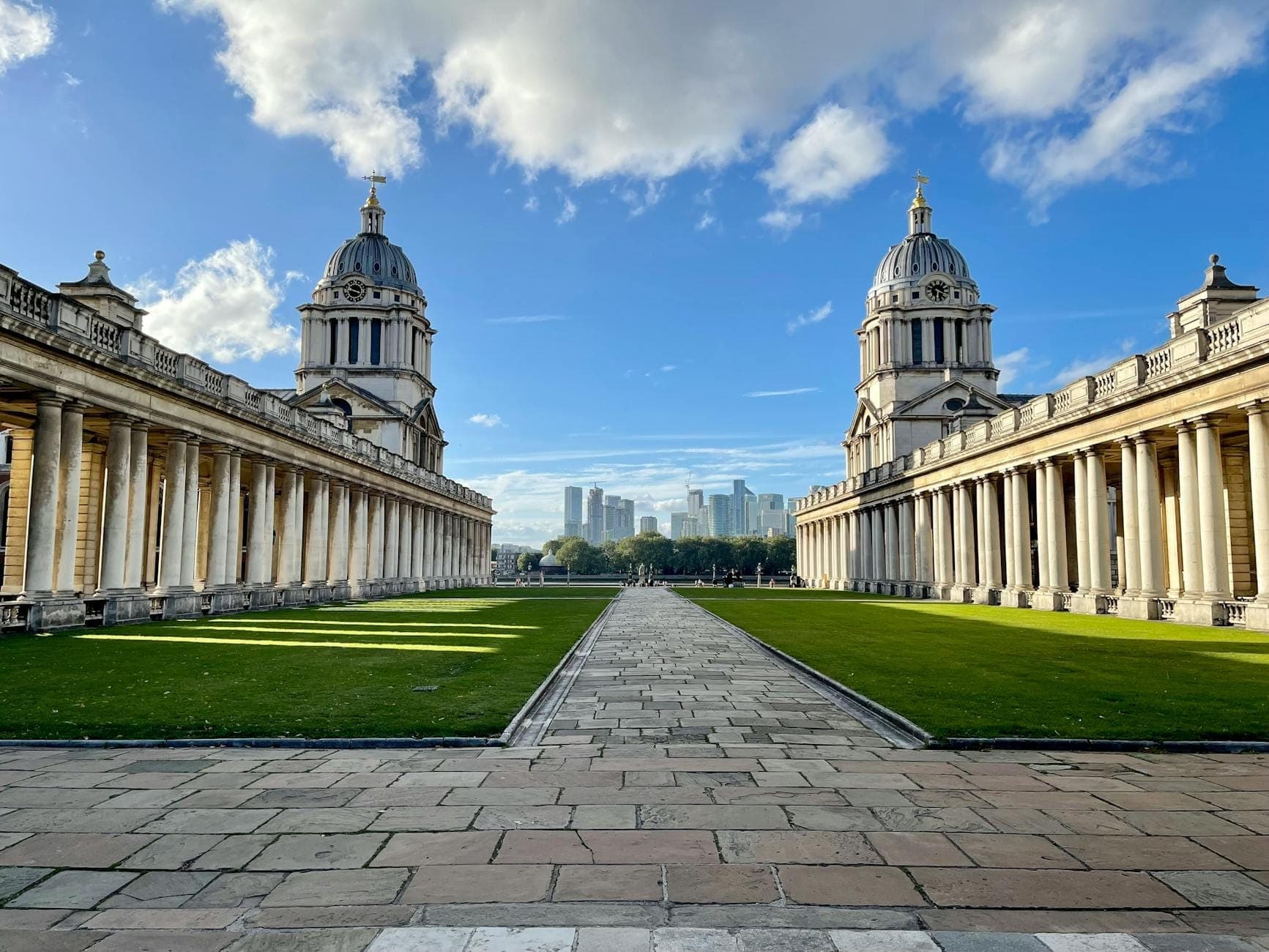 Blick auf das Old Royal Naval College mit den markanten Doppelkuppeln und den Kolonnaden unter einem dramatischen blauen Himmel.