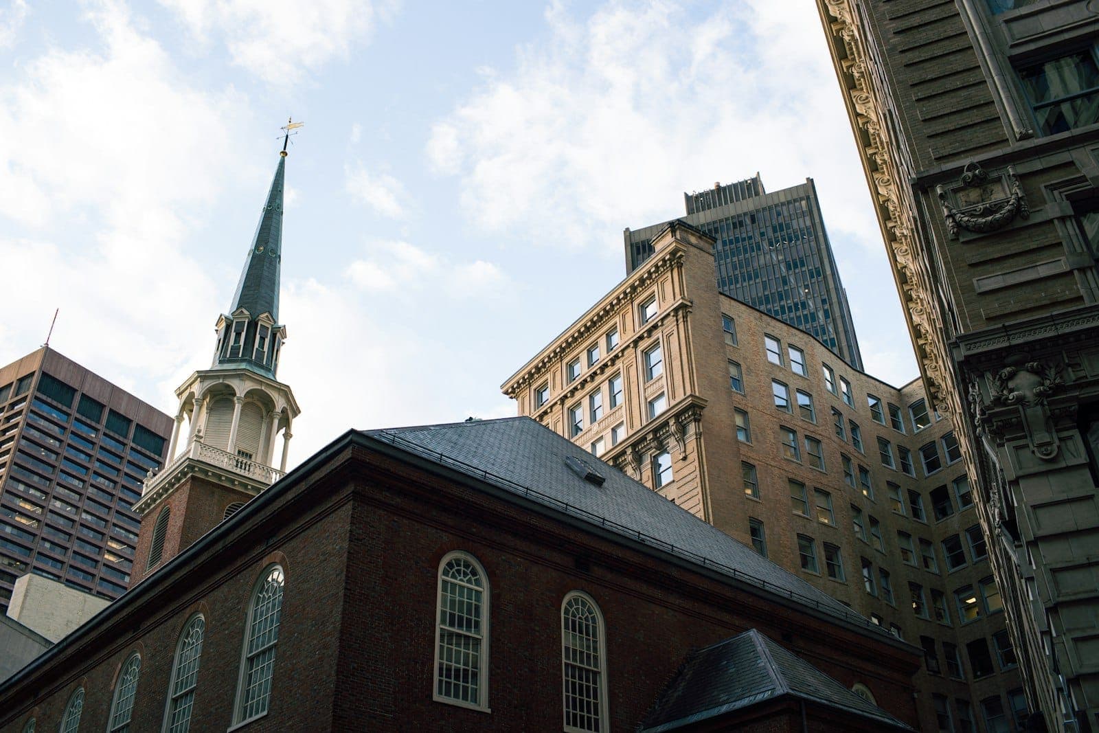 La façade en brique et le clocher de l'Old South Meeting House entourés de bâtiments modernes et historiques du centre-ville de Boston par une journée partiellement nuageuse.