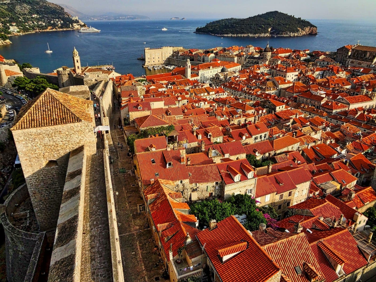 Ein weitläufiger Luftblick auf Dubrovniks Altstadt mit den charakteristischen Terrakotta-Dächern, den massiven mittelalterlichen Steinmauern und der Adria mit einer grünen Insel im Hintergrund im goldenen Sonnenlicht.