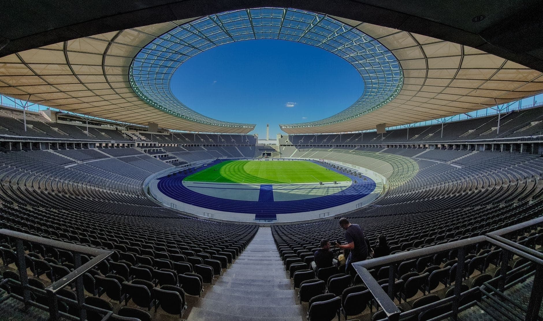 ภาพมุมกว้างจากอัฒจันทร์ใน Olympiastadion Berlin โชว์หลังคาทรงรีสมัยใหม่ ลู่วิ่งสีน้ำเงิน สนามหญ้าเขียวสด และแนวเก้าอี้ว่างยาวโค้งใต้ท้องฟ้าแจ่มใส