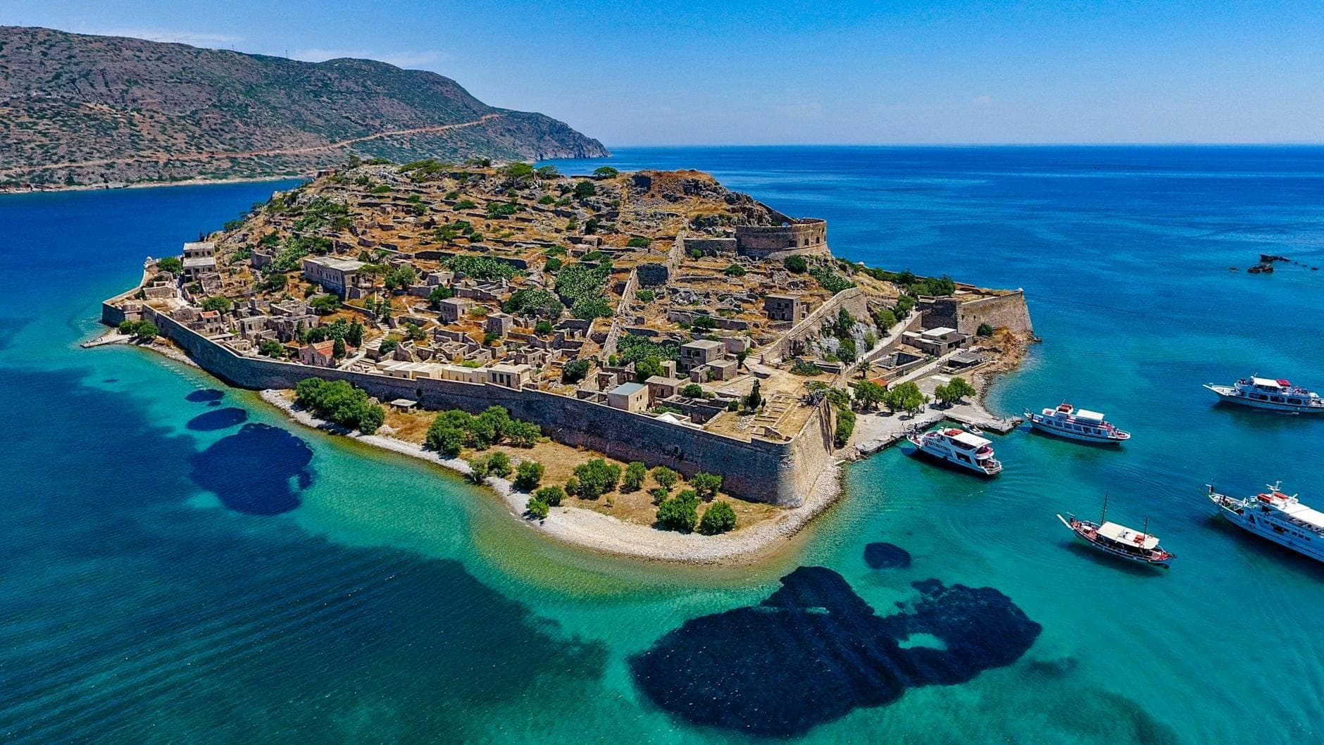 Vista aerea dell'isola di Spinalonga con le fortificazioni veneziane, acque turchesi e barche turistiche ancorate nelle vicinanze.