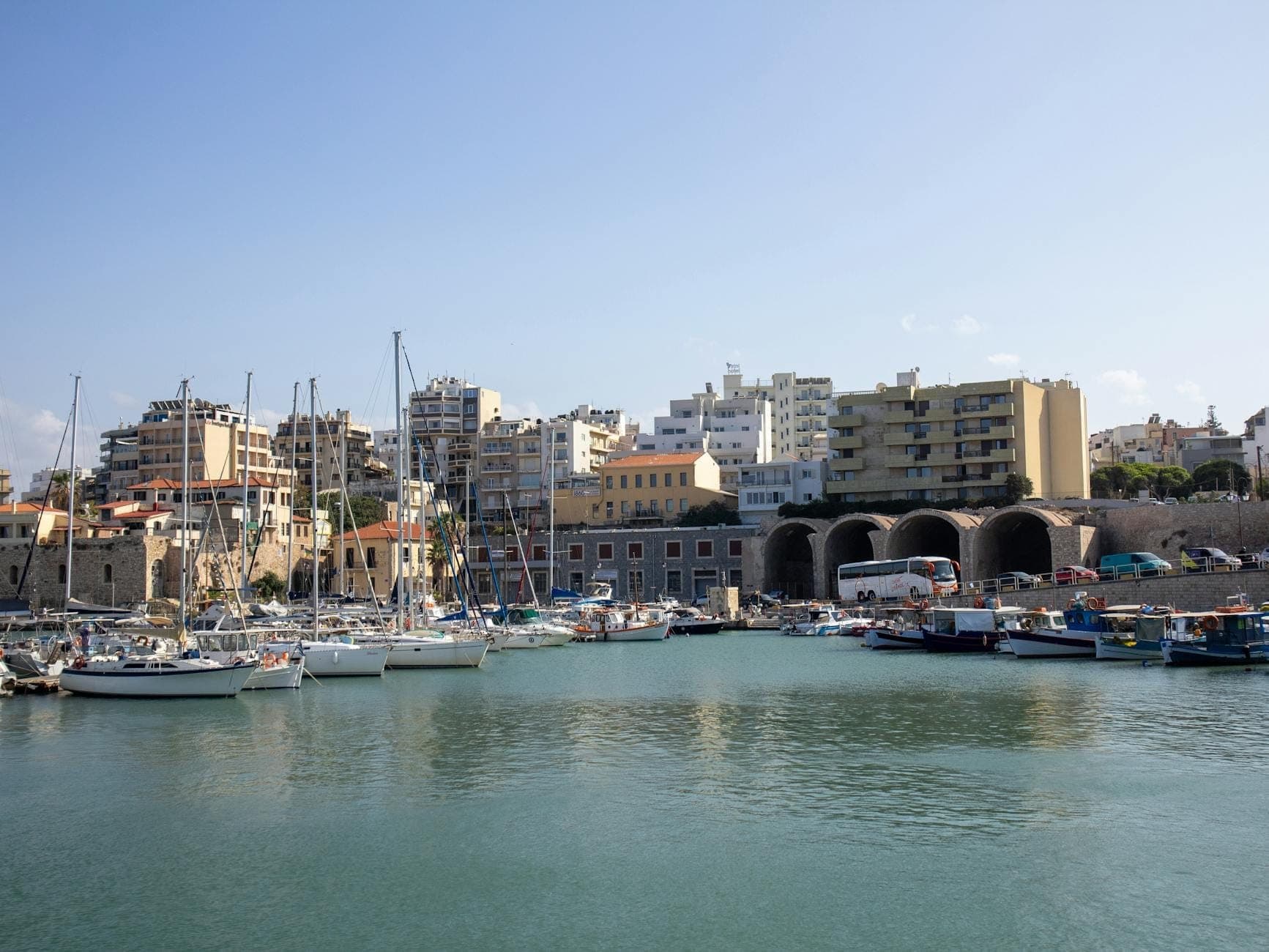 Yacht e barche da pesca ormeggiate nel porto di Heraklion, con la città e gli edifici sul lungomare sullo sfondo sotto un cielo sereno.