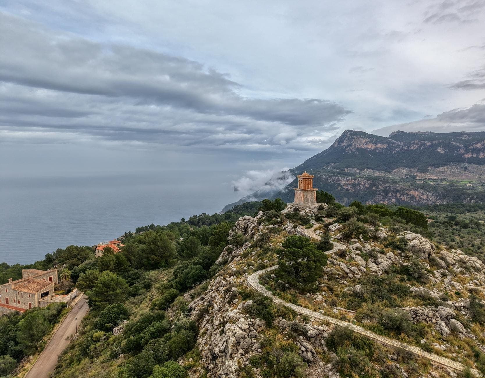 Steingebäude auf einem Hügel mit gewundenem Pfad, mit Blick auf die schroffen Berge und das Mittelmeer in der Serra de Tramuntana auf Mallorca.