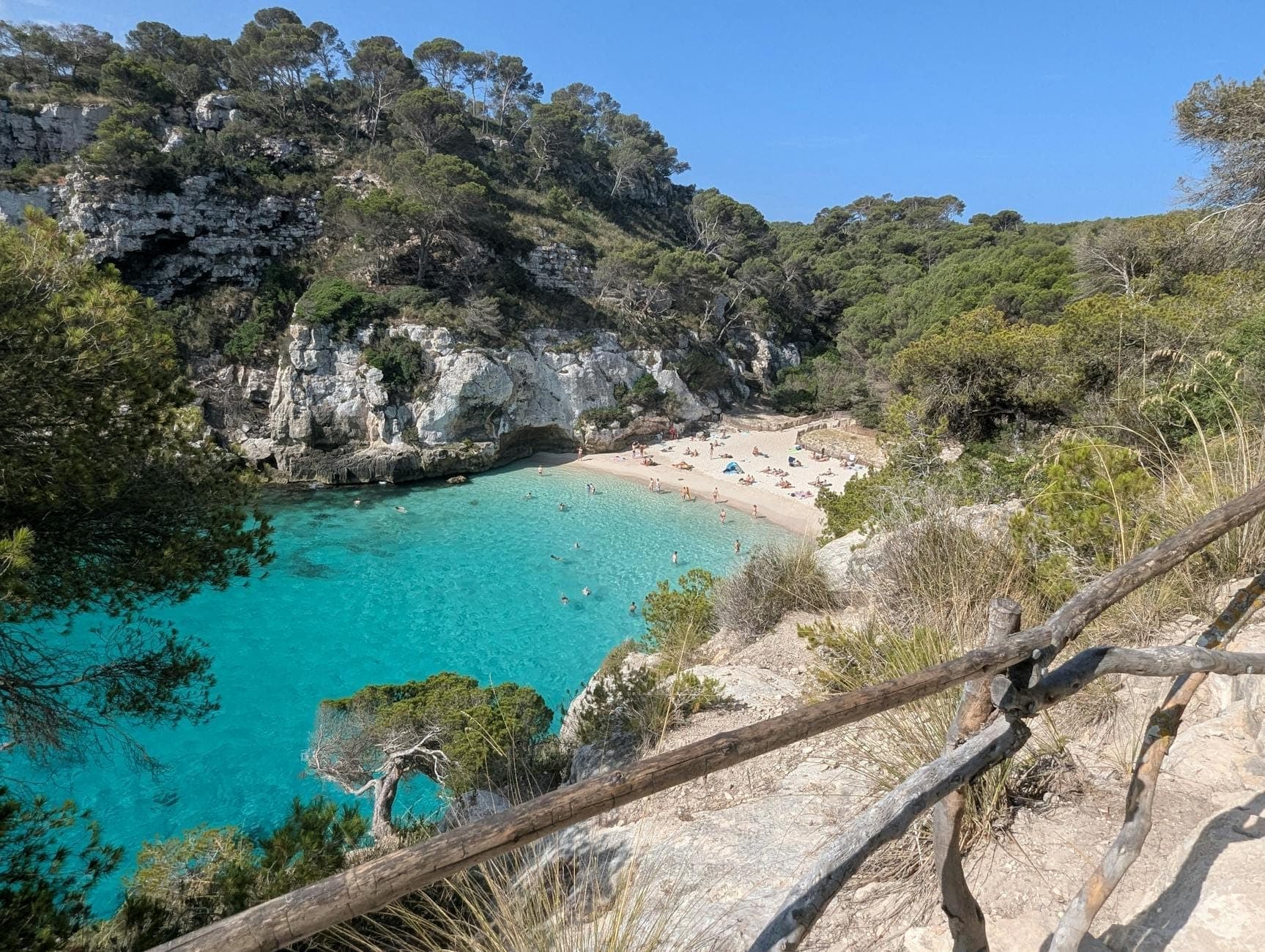 Kleine Bucht mit türkisfarbenem Wasser, Sandstrand, Sonnenbadenden und felsigen Klippen mit Pinien, von einem rustikalen Holzzaun aus gesehen.