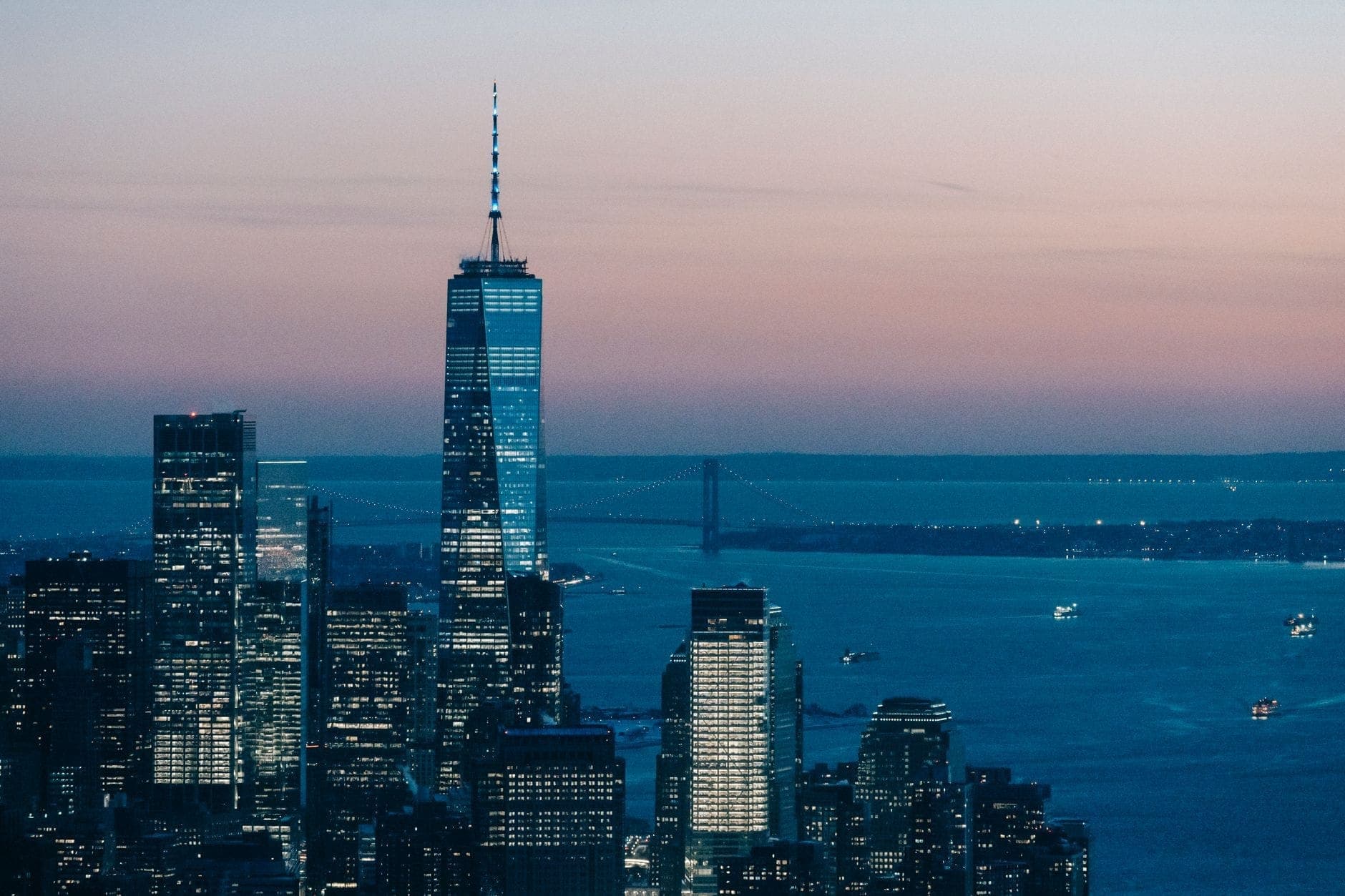 วิวของ One World Trade Center ที่สูงตระหง่านเหนือตึกระฟ้าใน Manhattan ยามพลบค่ำ พร้อมกับแม่น้ำ Hudson และสะพานที่อยู่ไกลออกไป