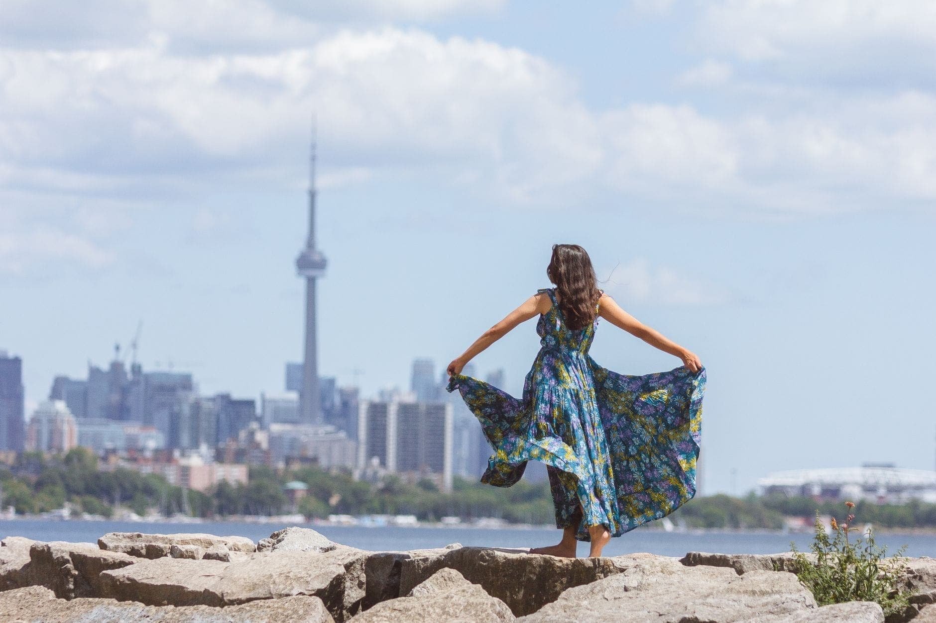 Donna con un vestito colorato in piedi sulla riva rocciosa di Ontario Place, con lo skyline di Toronto e la CN Tower visibili dall'altra parte del lago Ontario.