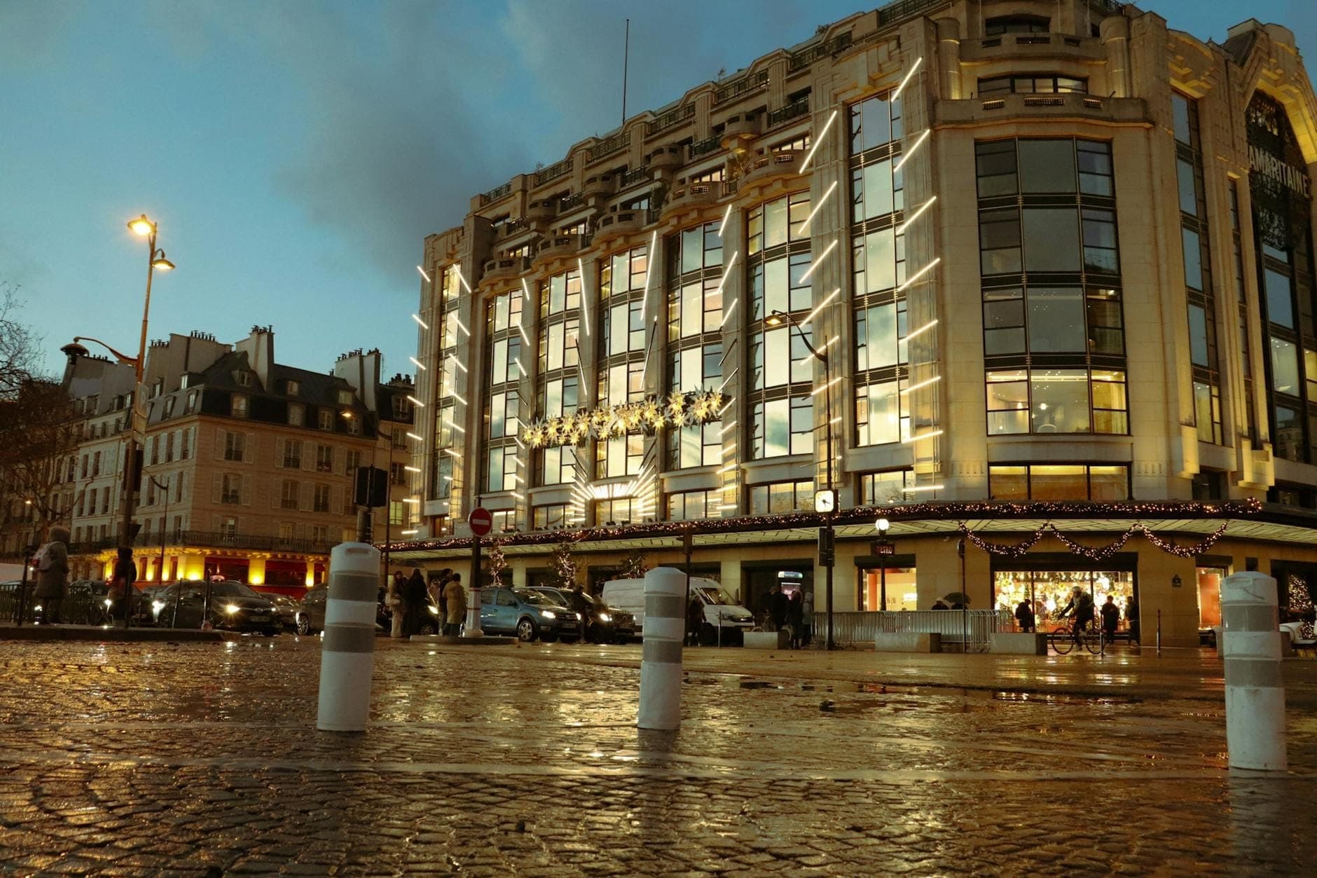 Modern glass and steel facade of Opéra Bastille illuminated at dusk, with people and cars in Place de la Bastille, Paris.