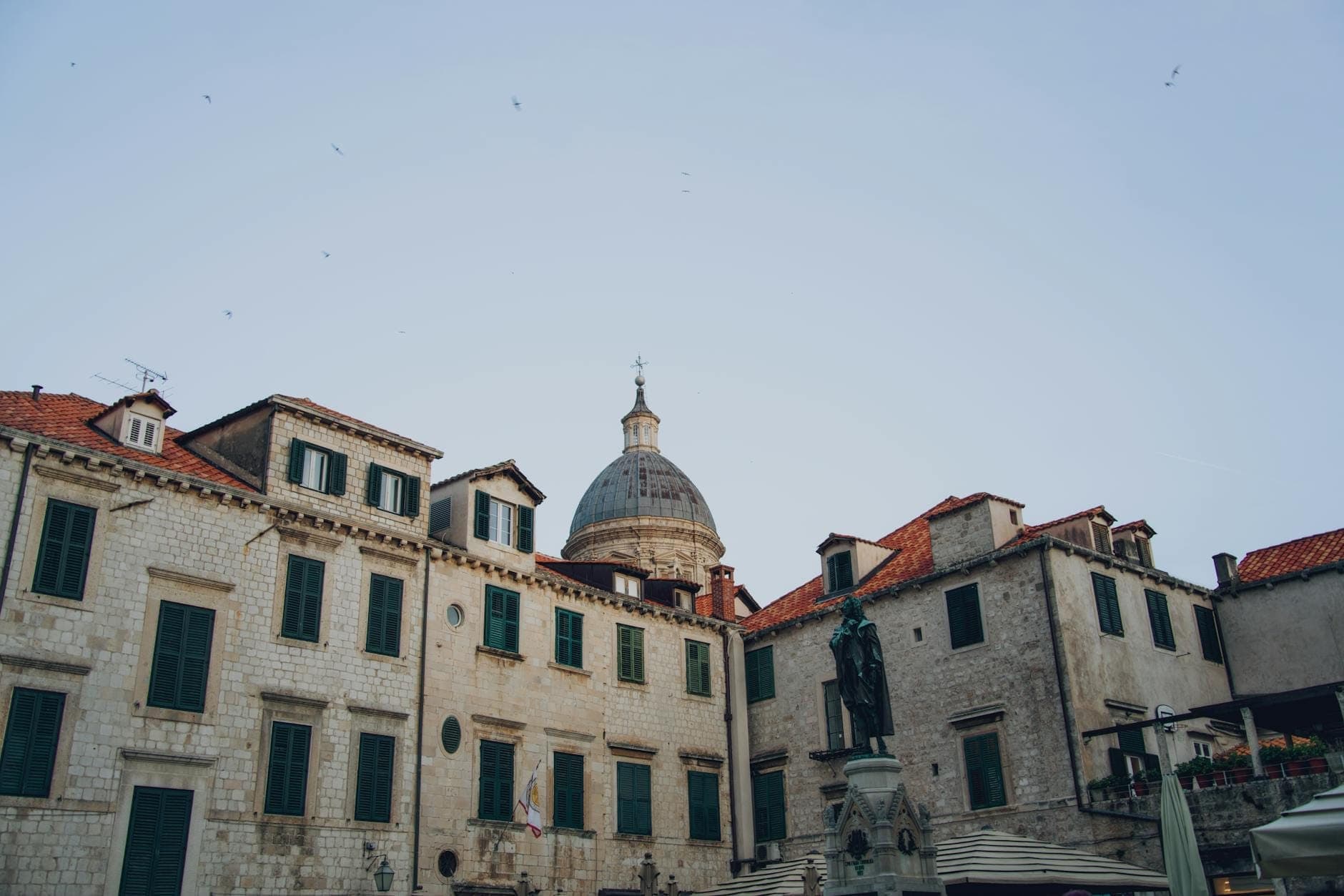 Vista da Praça Luža na Cidade Velha de Dubrovnik, com edifícios históricos de calcário, uma imponente igreja com cúpula e a Coluna de Orlando visível ao centro.
