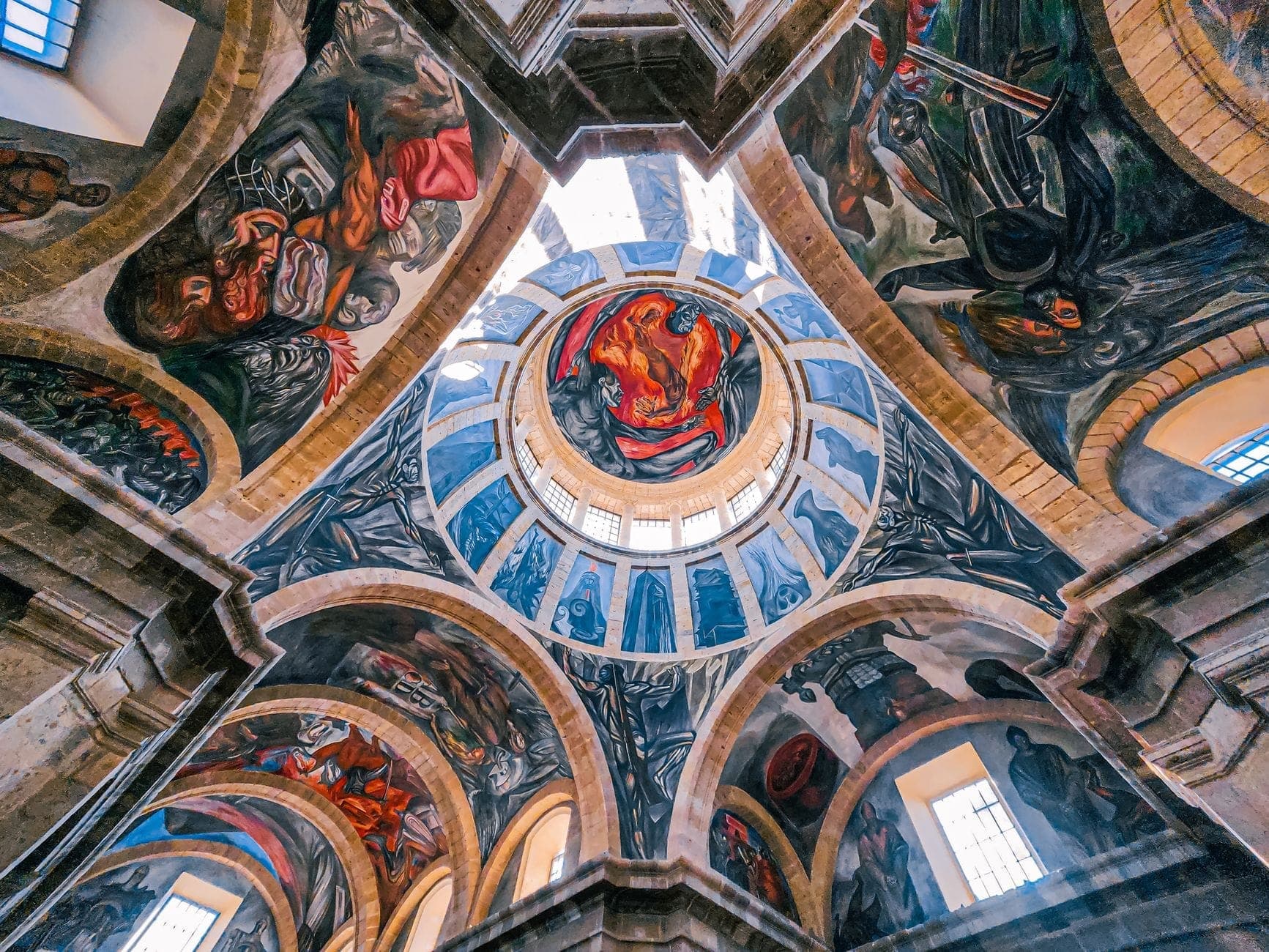ภาพมุมกว้างของ Hospicio Cabañas ภายในอาคาร โดดเด่นด้วยจิตรกรรมฝาผนังสีสดใสของโอรอสโกทั้งบนโดมและซุ้มโค้ง ตัดกับแสงธรรมชาติที่ส่องผ่านหน้าต่าง