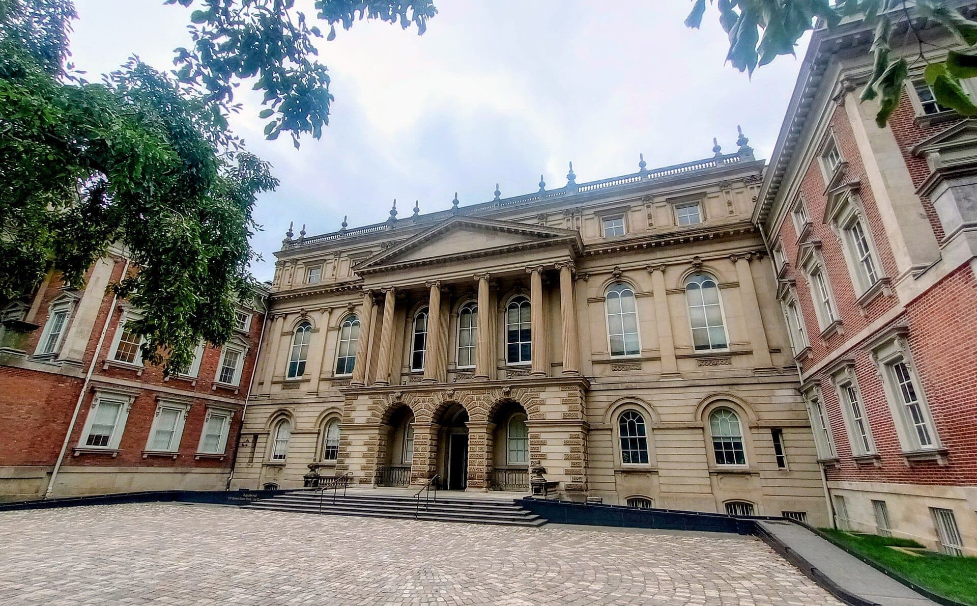 Vista frontale della grandiosa facciata neoclassica dell'Osgoode Hall con colonne in pietra, ingresso ad arco e ali in mattoni rossi in una giornata nuvolosa.